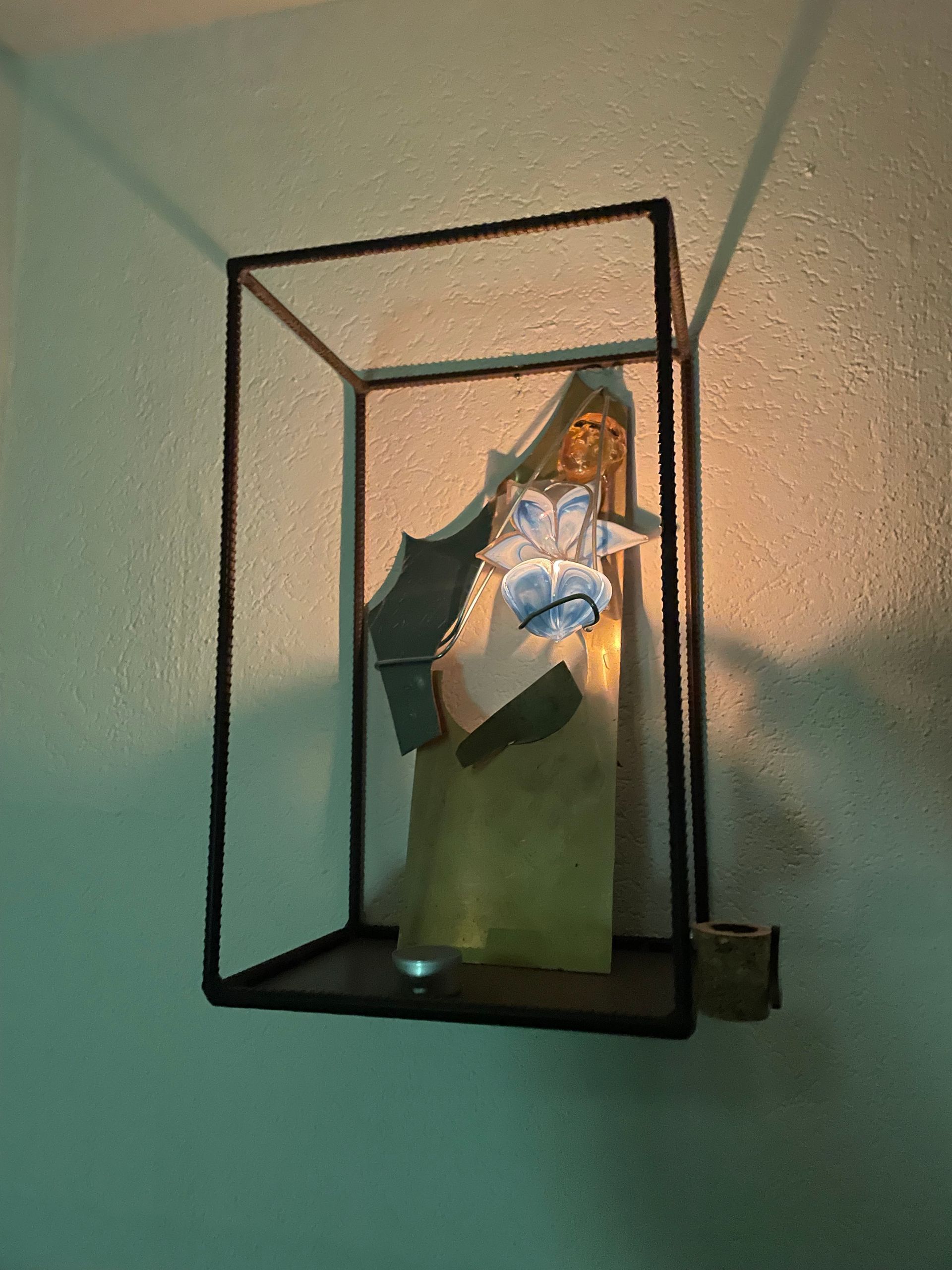 Een kunstwerk met een metalen frame en verlichte glasstukken erin, gemonteerd aan een turquoise muur.