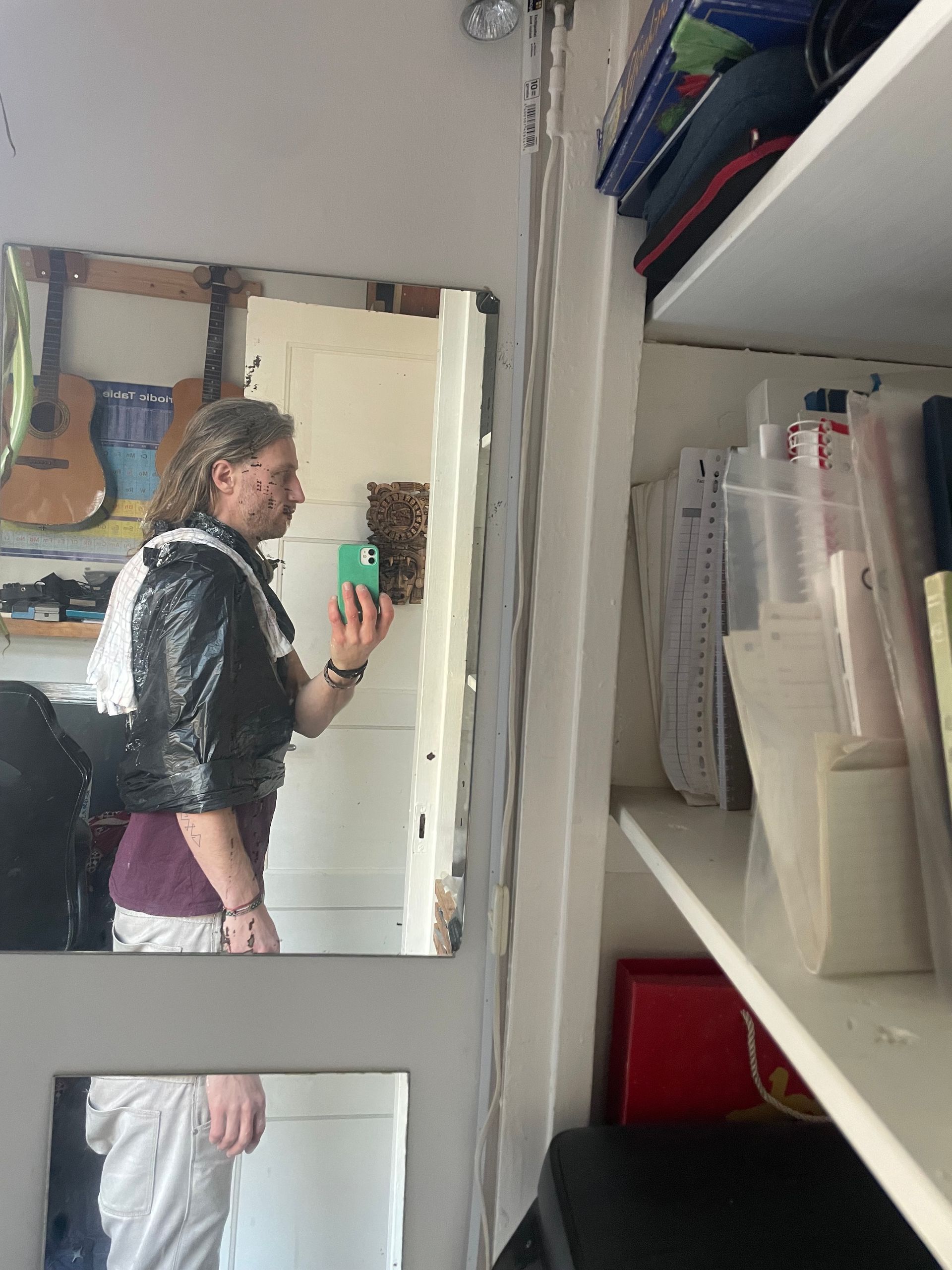 Een persoon in een jasje met pailletten maakt een selfie in de spiegel. Rechts daarvan staat een plank met kantoorartikelen.
