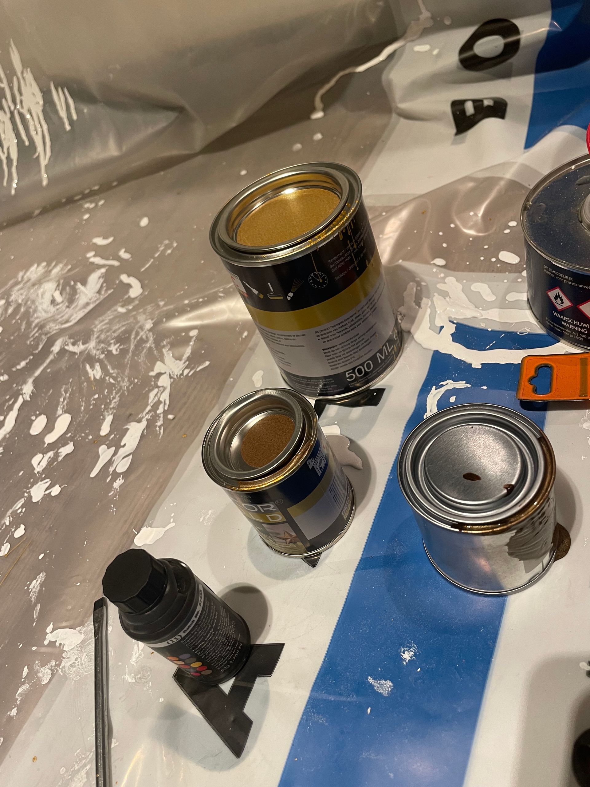 Verfblikken, waarvan één open, op een wit-blauwe ondergrond met verfspatten en een zwarte fles.