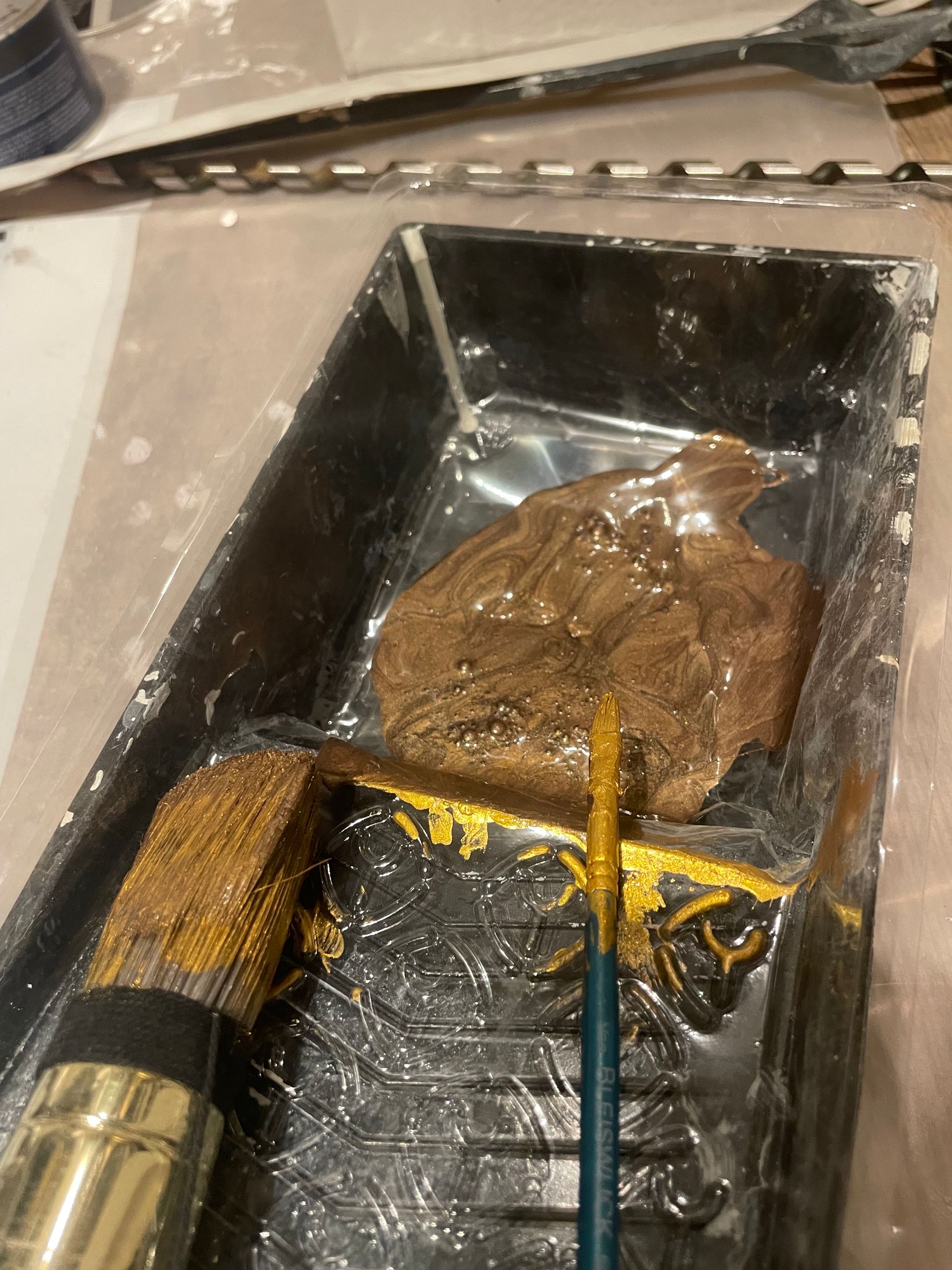Verfbakje met bruine verf en twee kwasten, een gouden en een blauwe.