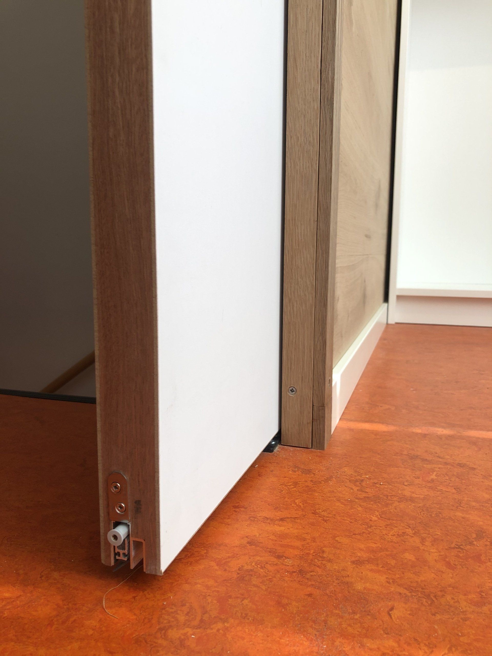 Een witte deur staat op een kier naast een licht houten muur. De oranje vloer is zichtbaar.
