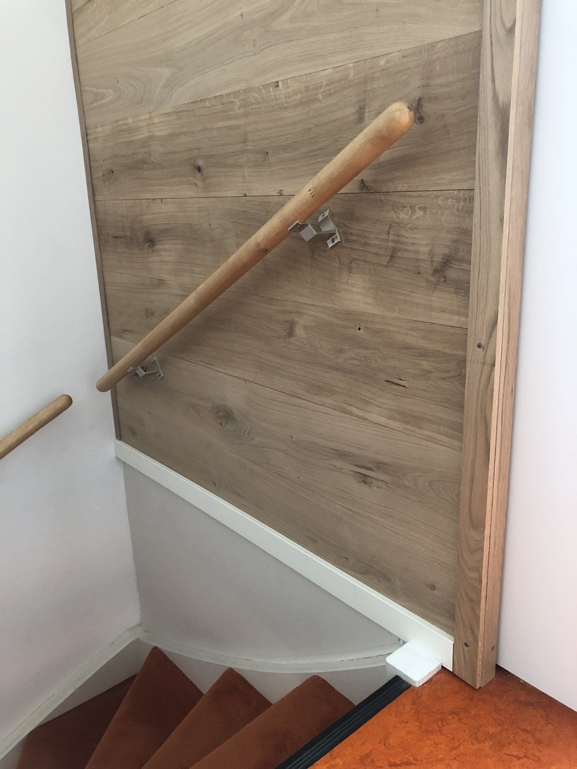 Houten trap met leuning. Bruine vloerbedekking op de treden. Beige muur en houten lambrisering.