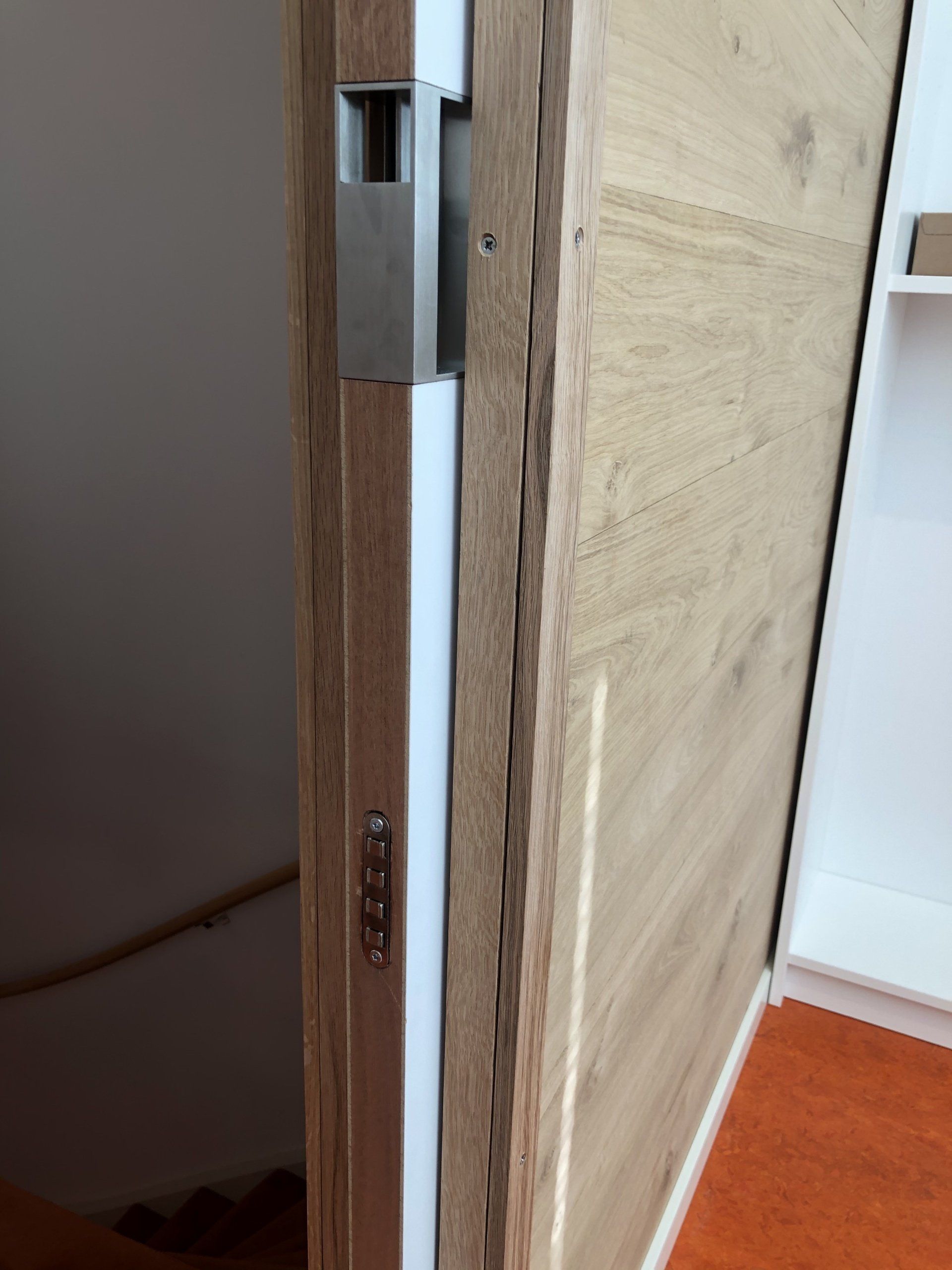 Houten deur met roestvrijstalen sluiting, witte omlijsting en een glimp van een trap op de achtergrond.