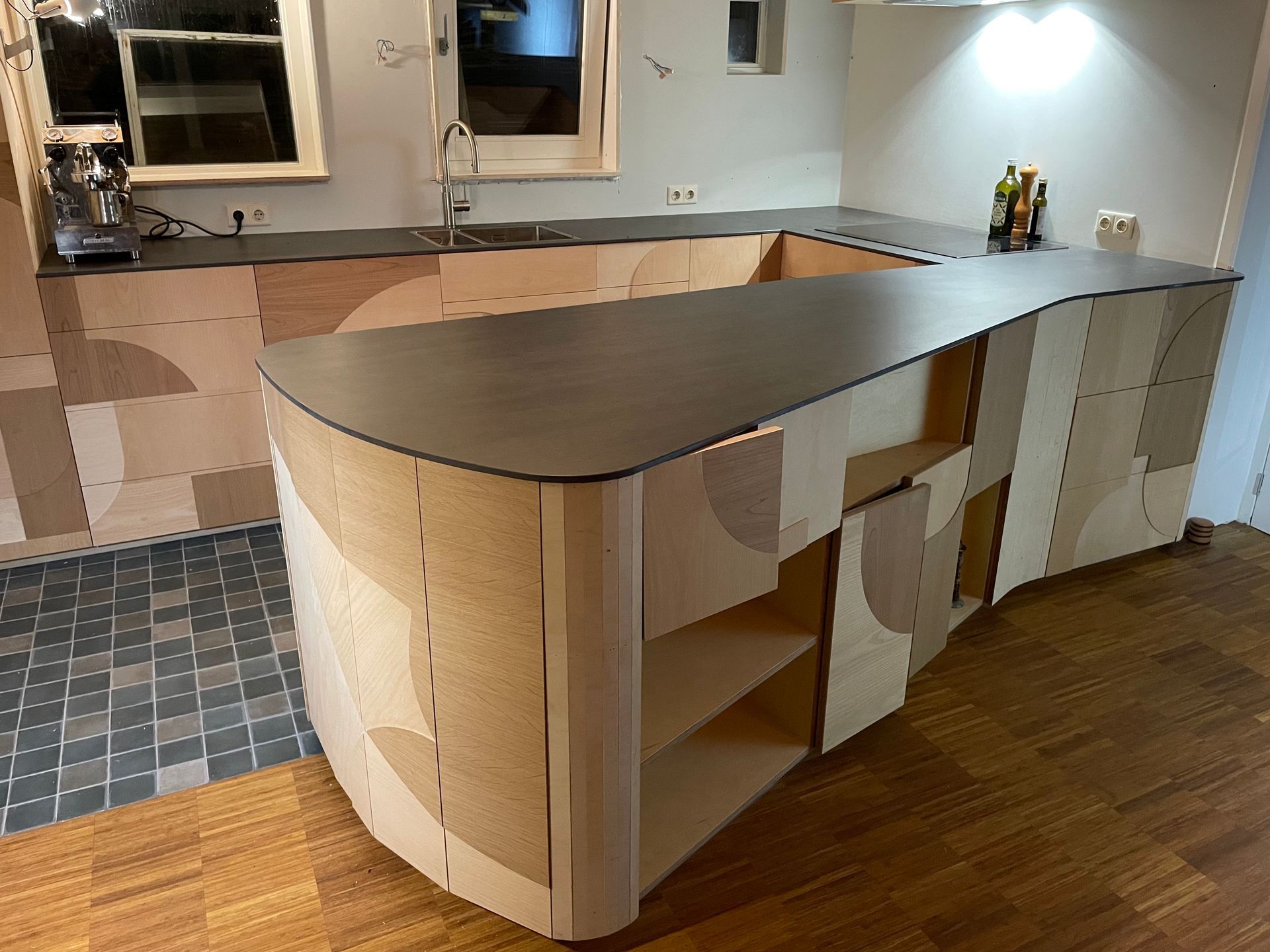 Op maat gemaakte houten keuken met gebogen kookeiland en donker aanrechtblad. Lichte houten kasten met geometrisch design.