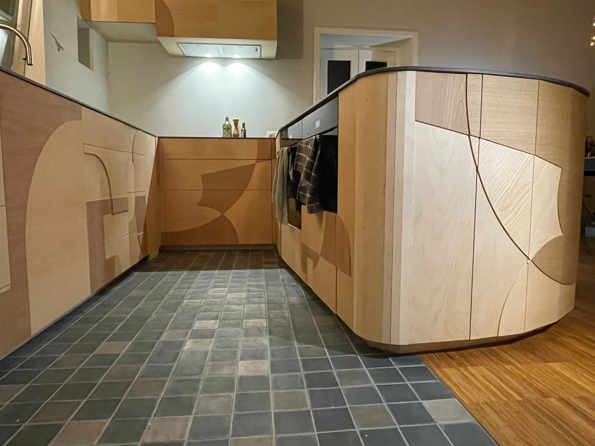 Moderne keuken met gebogen houten kasten en geometrische vormen; donkere tegelvloer.