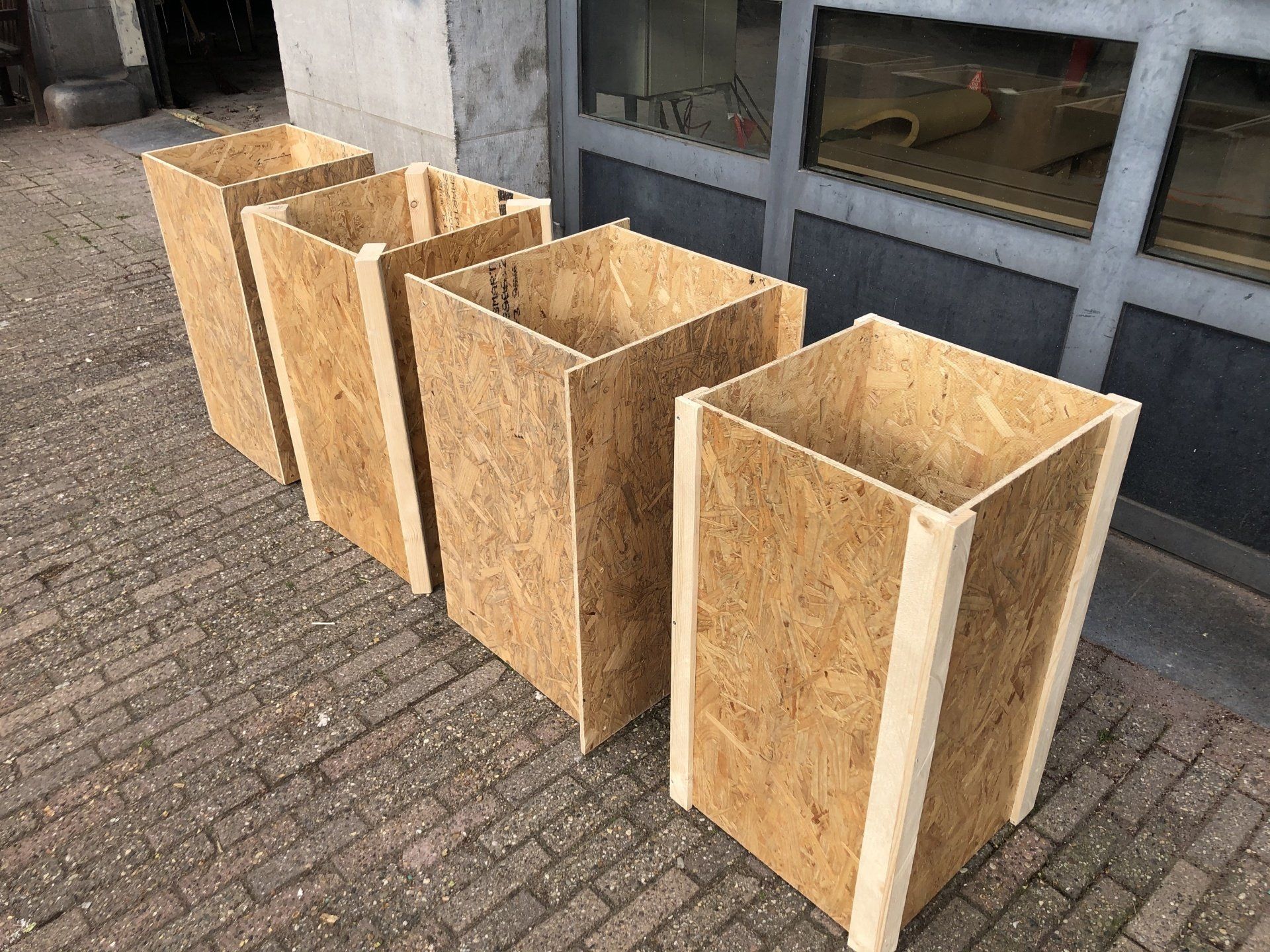 Vier rechthoekige houten plantenbakken van OSB, op een bakstenen ondergrond.