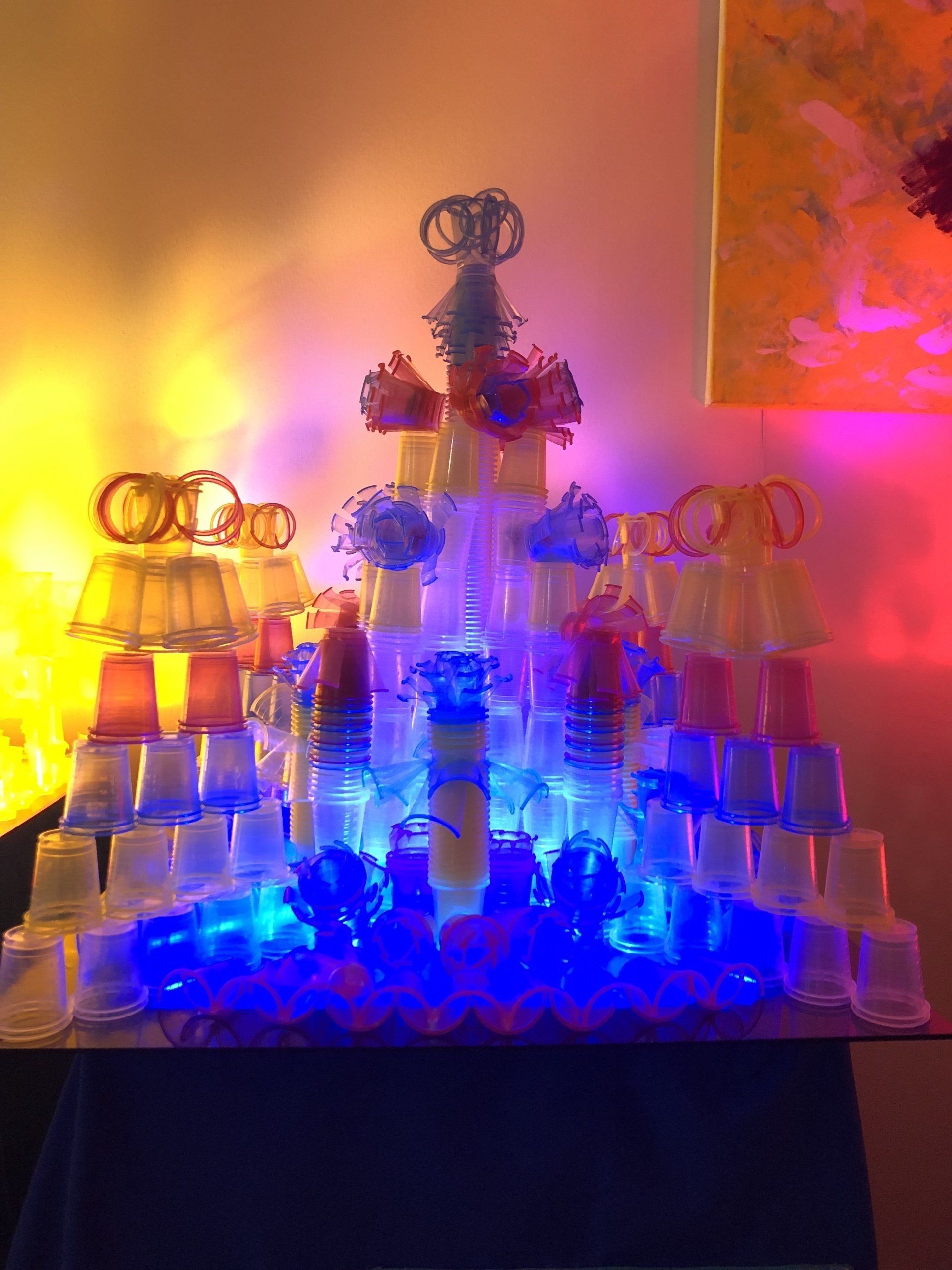 Piramidesculptuur gemaakt van plastic bekertjes, verlicht met blauwe, gele en rode lampen.