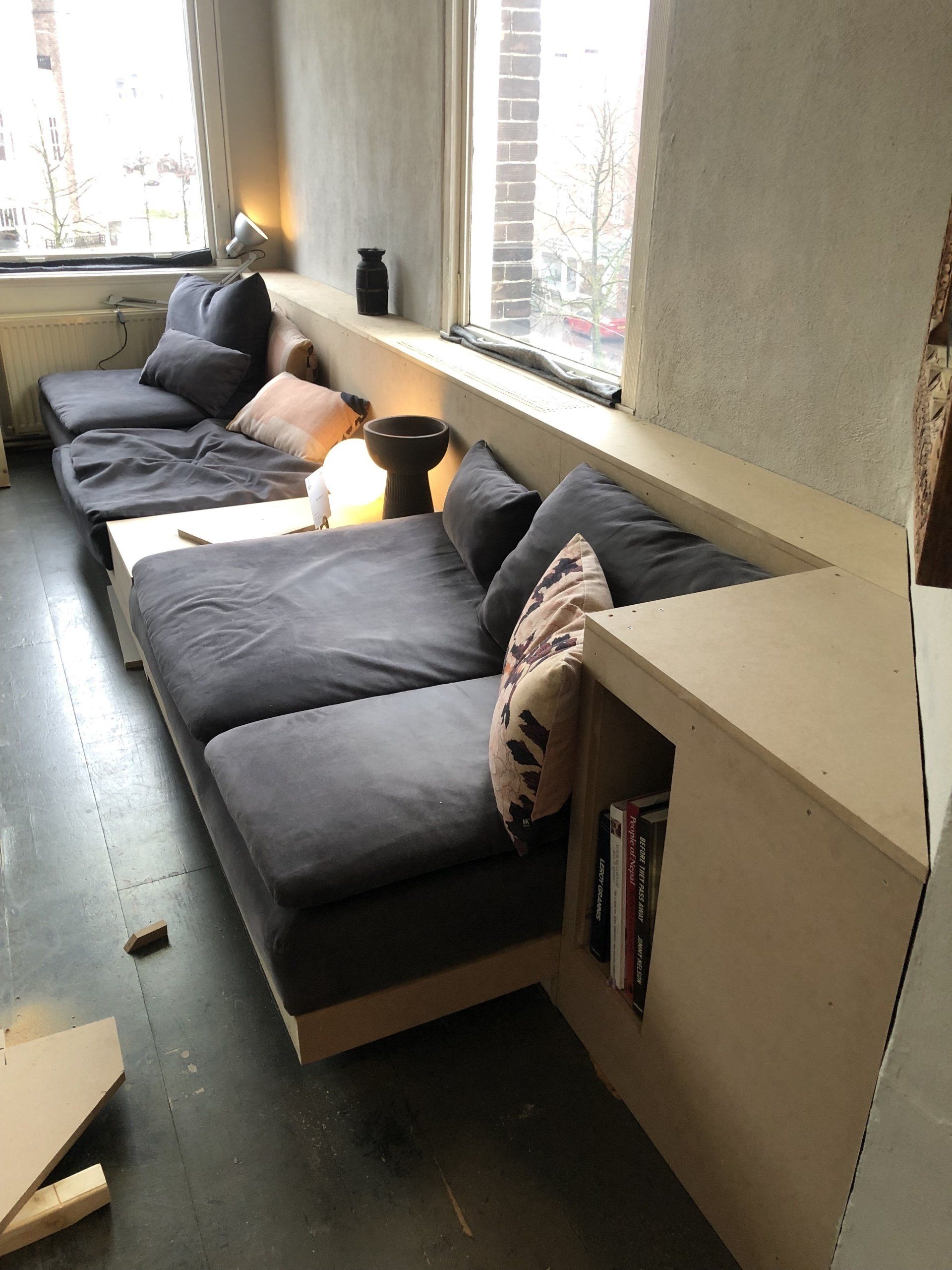 Een ingebouwde grijze hoekbank met een plank en een kleine boekenkast onder een raam.