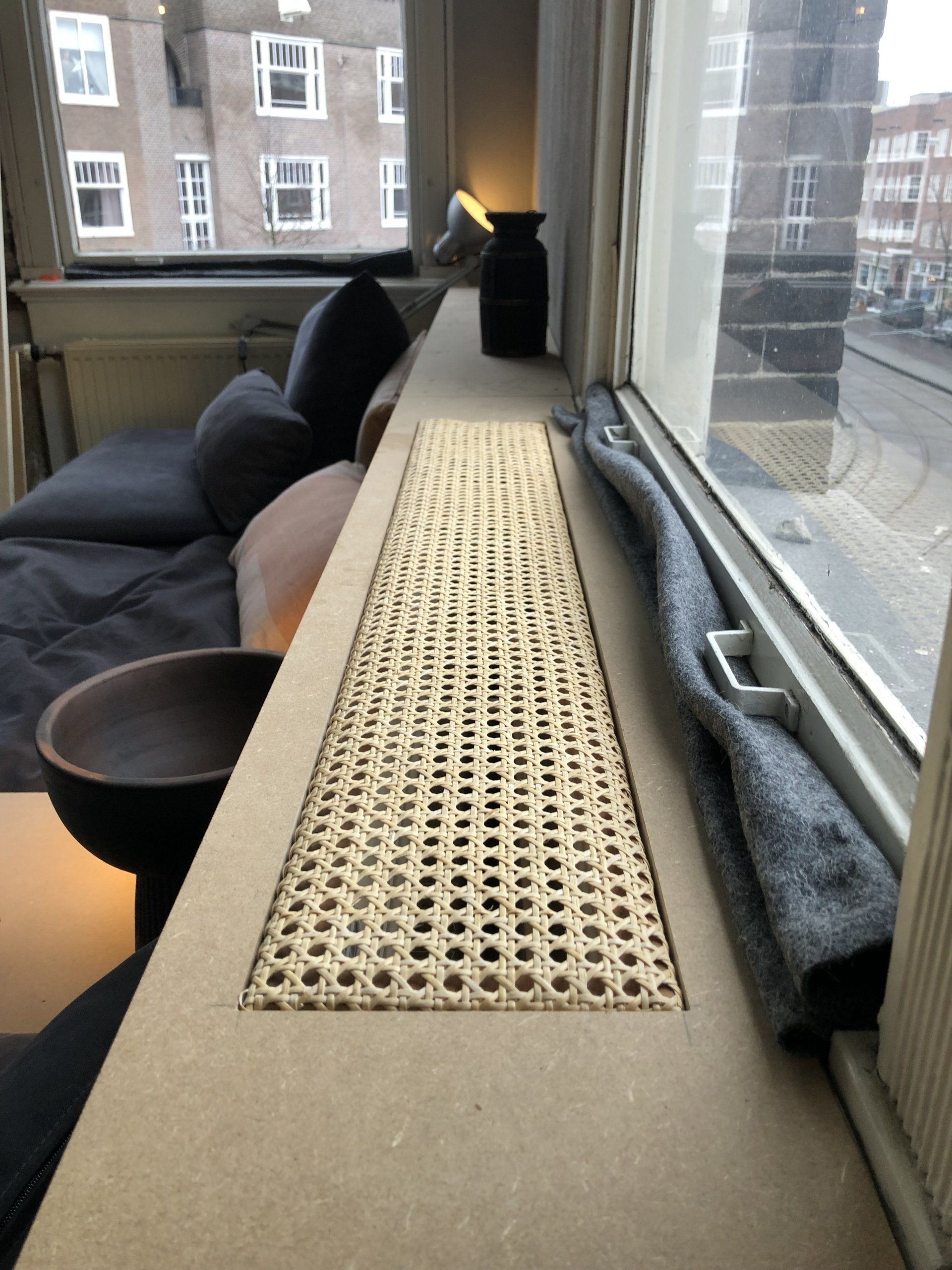 Vensterbank met radiatorbekleding van geweven riet, met uitzicht op een stadsstraat.
