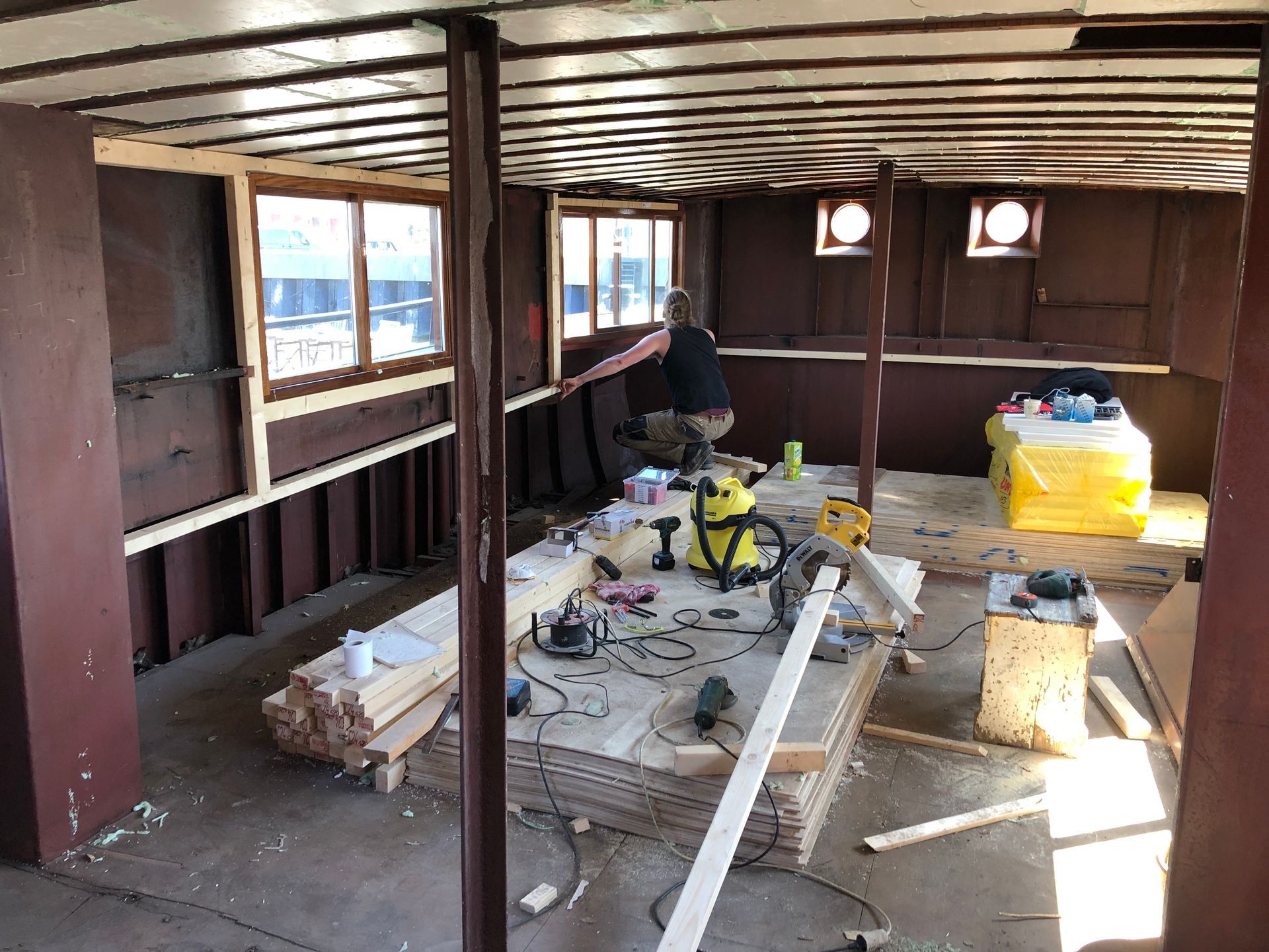 Interieur van een ruimte in aanbouw; iemand werkt met hout, gereedschap ligt verspreid over een multiplex ondergrond.