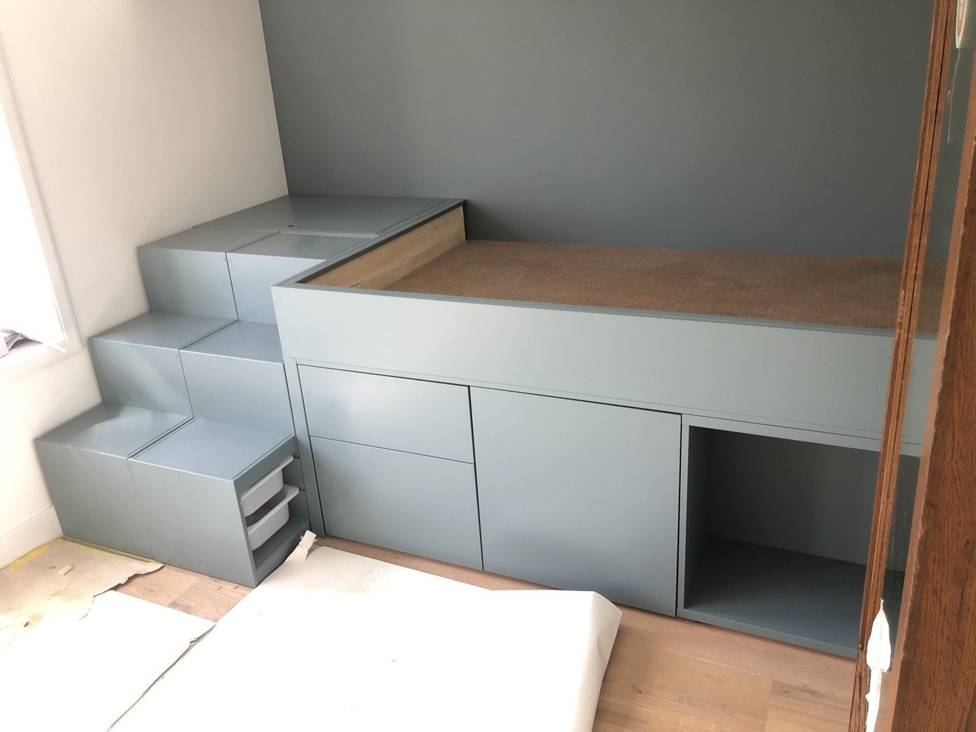 Ingebouwd bed met opbergruimte. Grijze treden en kastjes leiden naar het bedplatform tegen een blauwe muur.