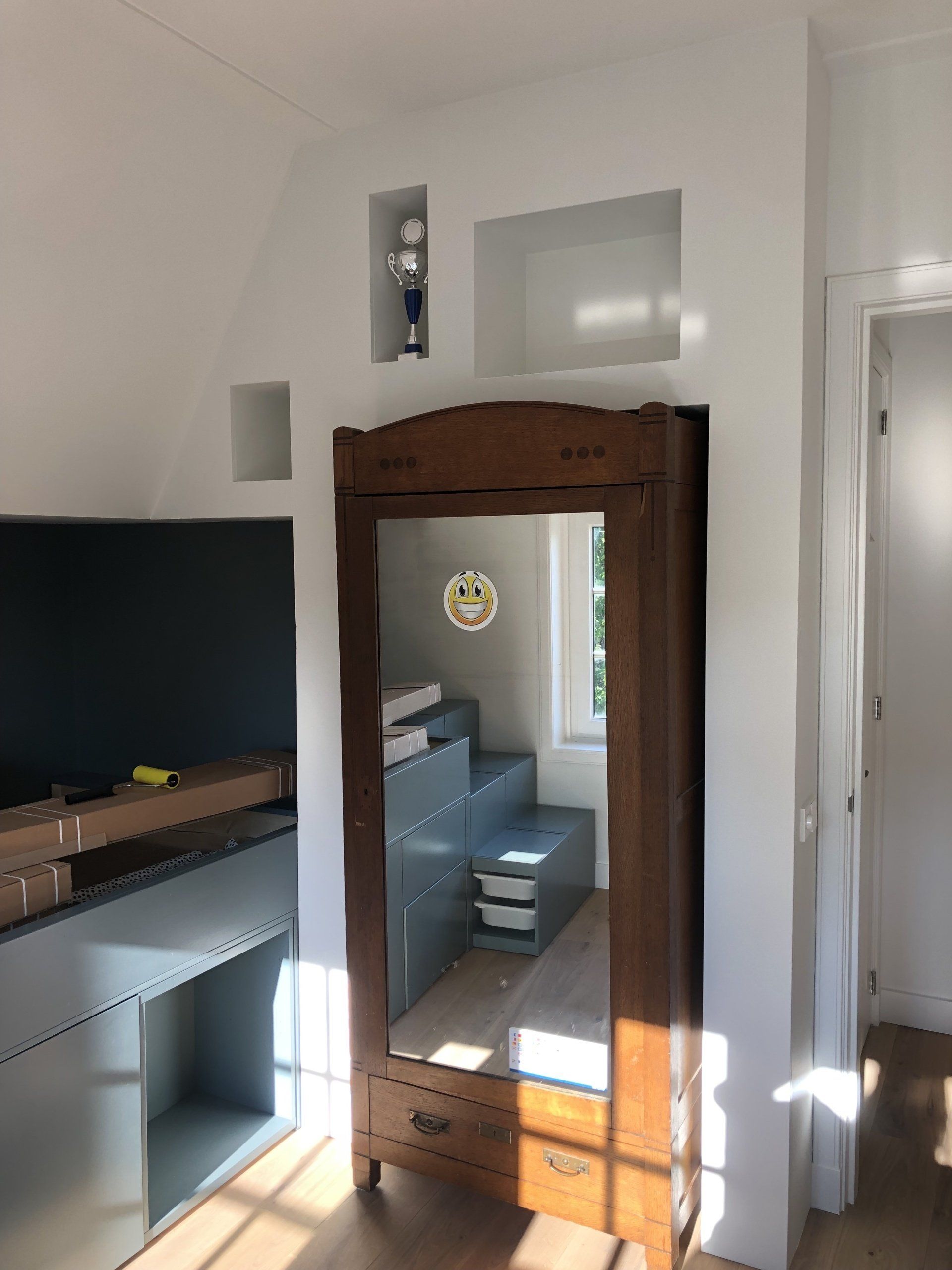 Houten kledingkast met spiegel, ingebouwde planken in een witte muur en een glimp van een kleine kamer.