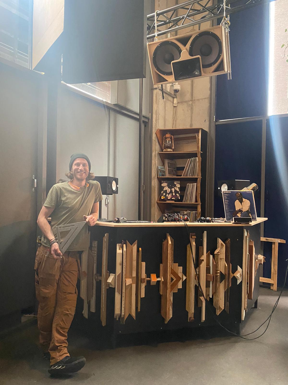 Een persoon glimlacht naast een op maat gemaakte dj-booth met houten details en hoge luidsprekers erboven, in een studio.