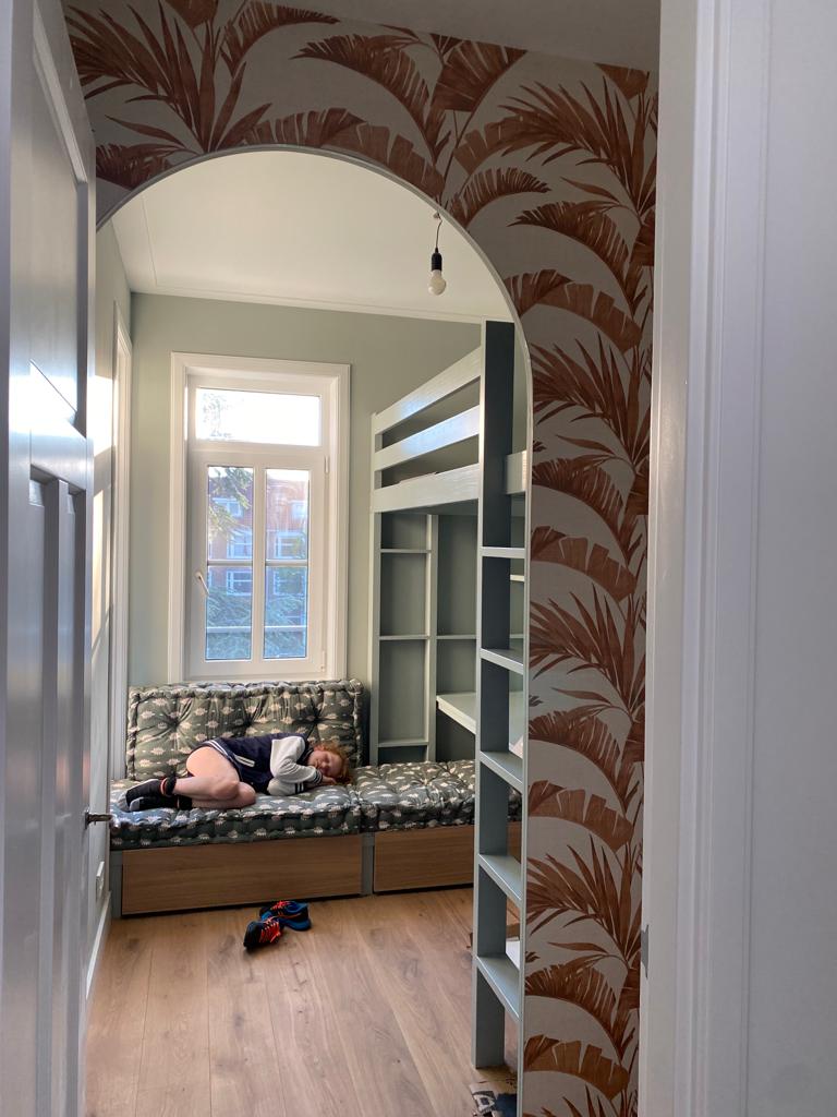 Kinderkamer met een boogvormige deuropening, behang met palmbladeren, een stapelbed en een kind dat op een bank ligt.