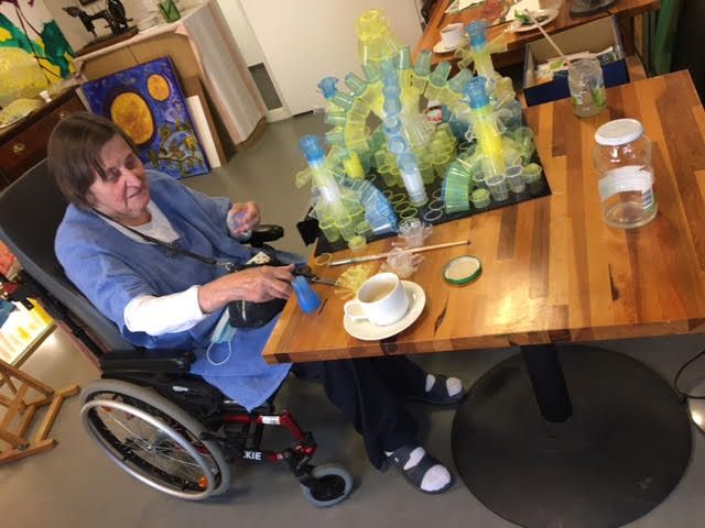 Vrouw in een rolstoel aan een tafel met een glazen kunstwerk, koffie en kunstbenodigdheden.