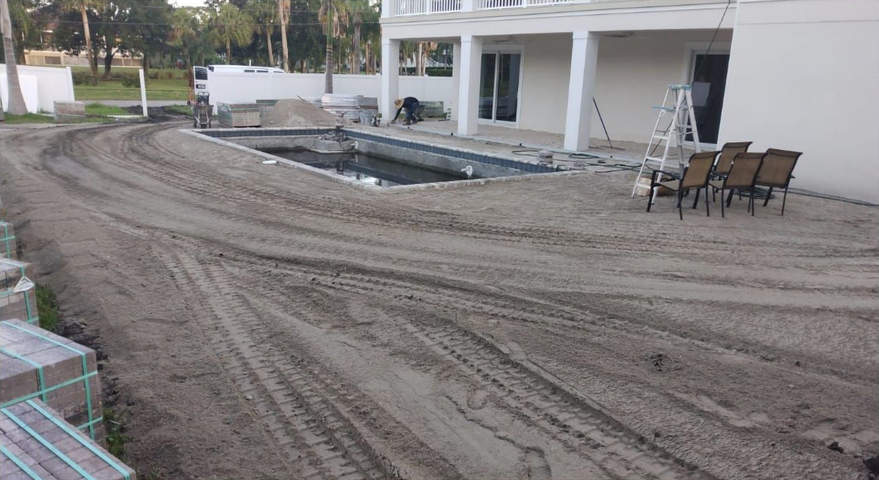 Construction site with a partially built pool, dirt, bricks, and a house.