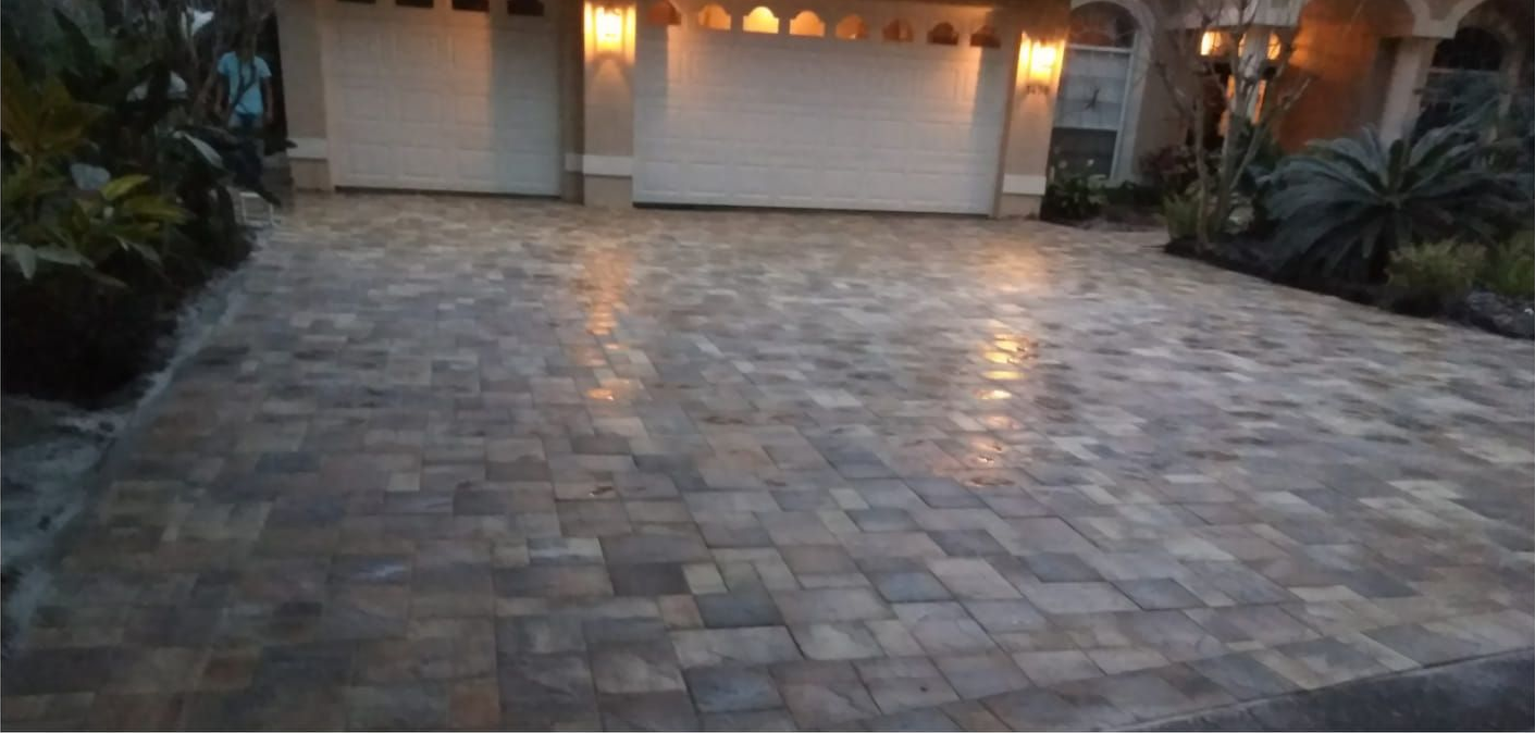 Driveway paved with textured, rectangular stones leading to a house with two garage doors lit by warm-toned lights.