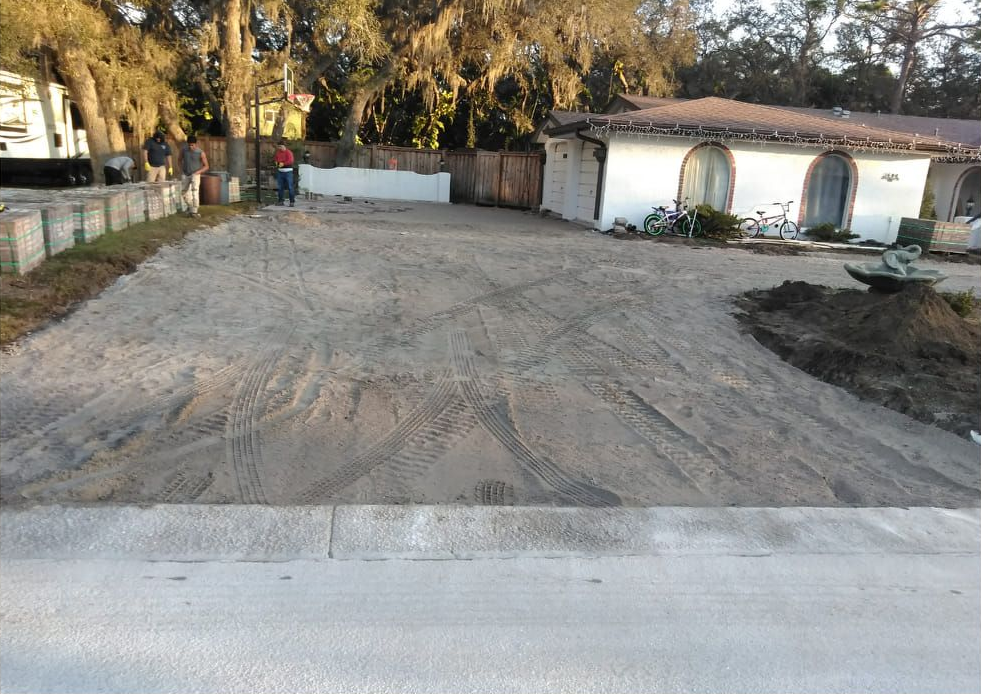 Driveway with dirt, house on the right, and people near a white structure.