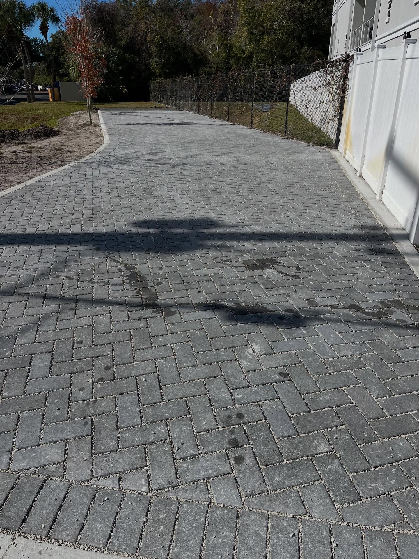 Brick driveway with gray pavers, leading toward trees and a building with white panels.