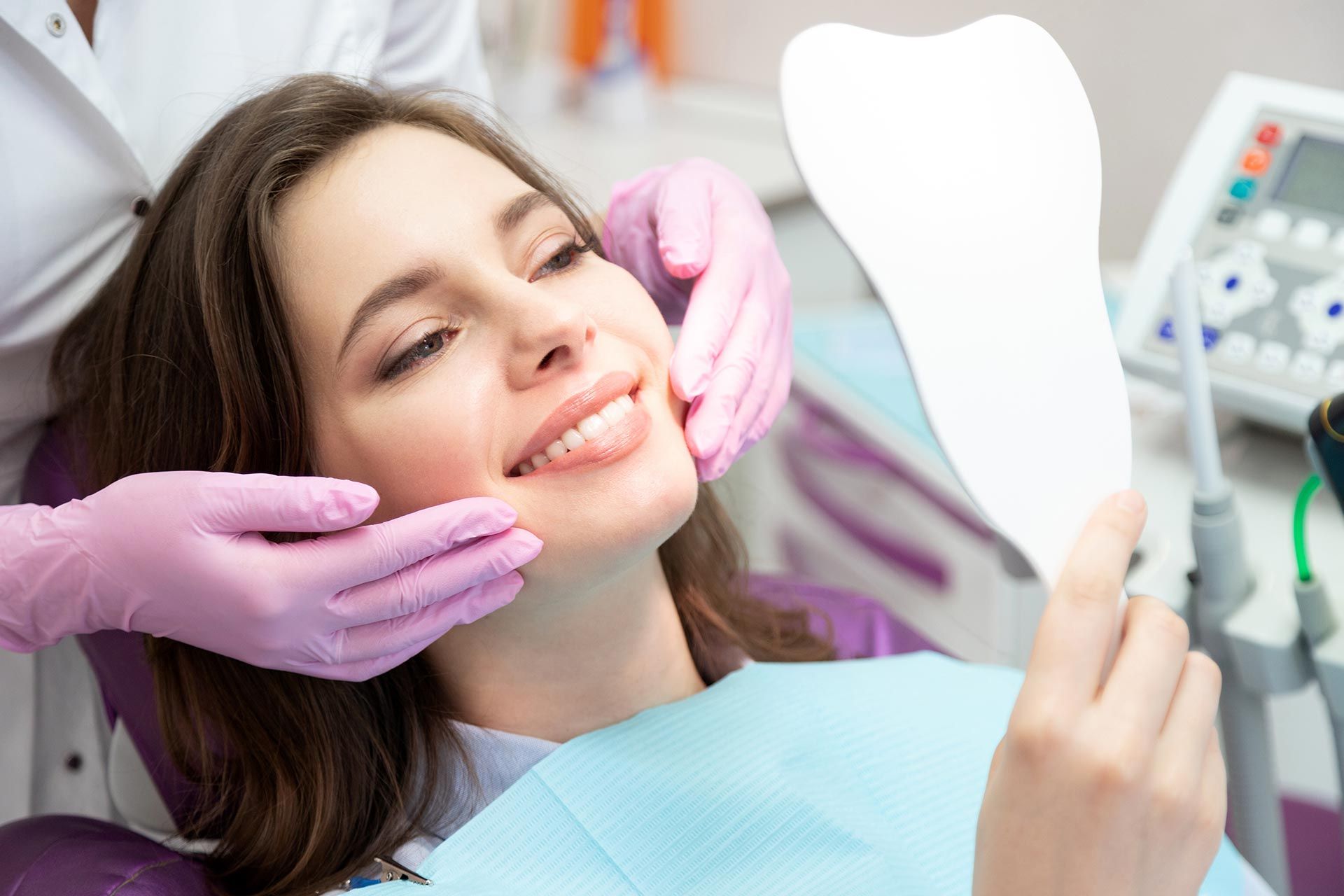 Woman in a dental chair smiling while gloved hands adjust her face, with dental tools nearby.