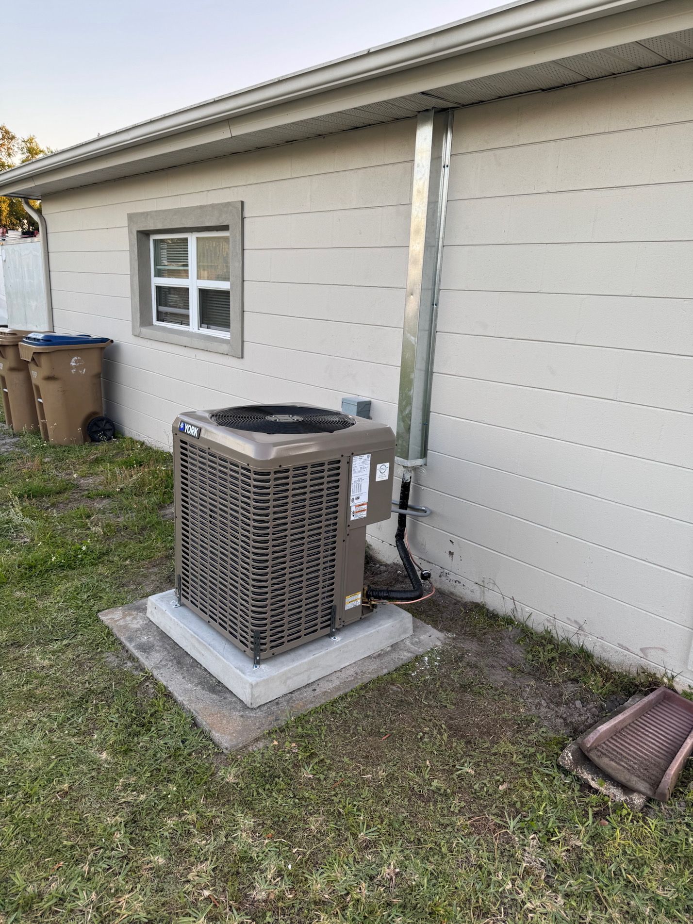 Outdoor HVAC unit beside a beige house wall, near a small window and grass yard