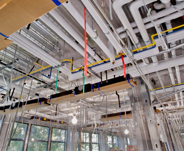 Construction site ceiling with exposed pipes, ducts, and hanging cables above large windows