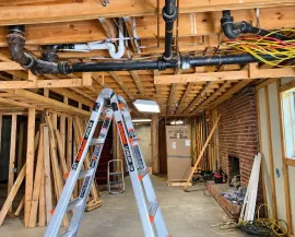 Ladder in unfinished basement with exposed ceiling beams, pipes, wiring, and renovation materials.