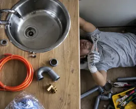 Plumber working under a sink beside tools, pipes, and orange tubing on a wooden floor