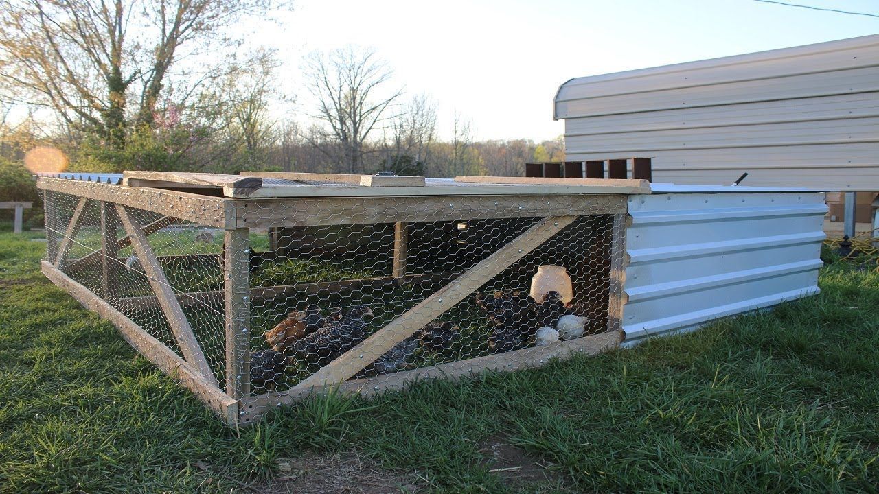 A low, wooden-framed chicken coop with wire mesh siding sits on a grassy field, featuring a white metal enclosed area.