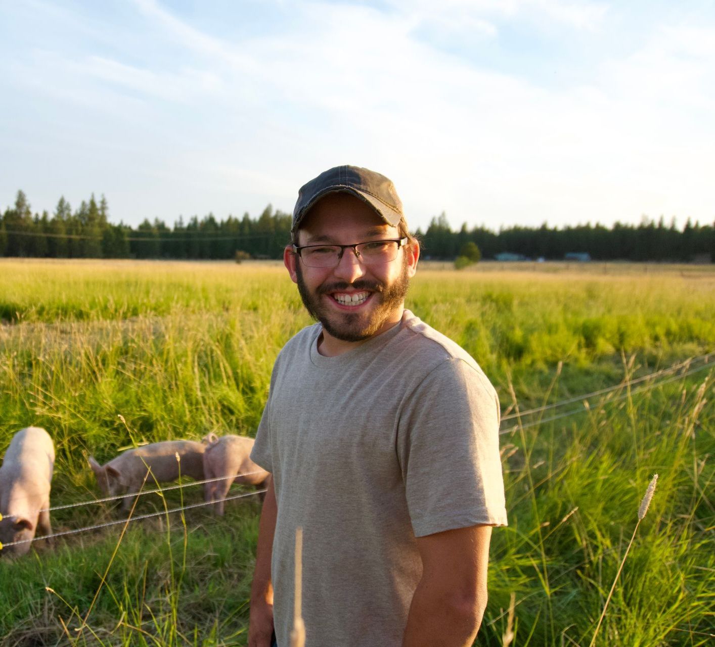 A smiling person in a hat and glasses stands in a grassy field with pigs in the background.