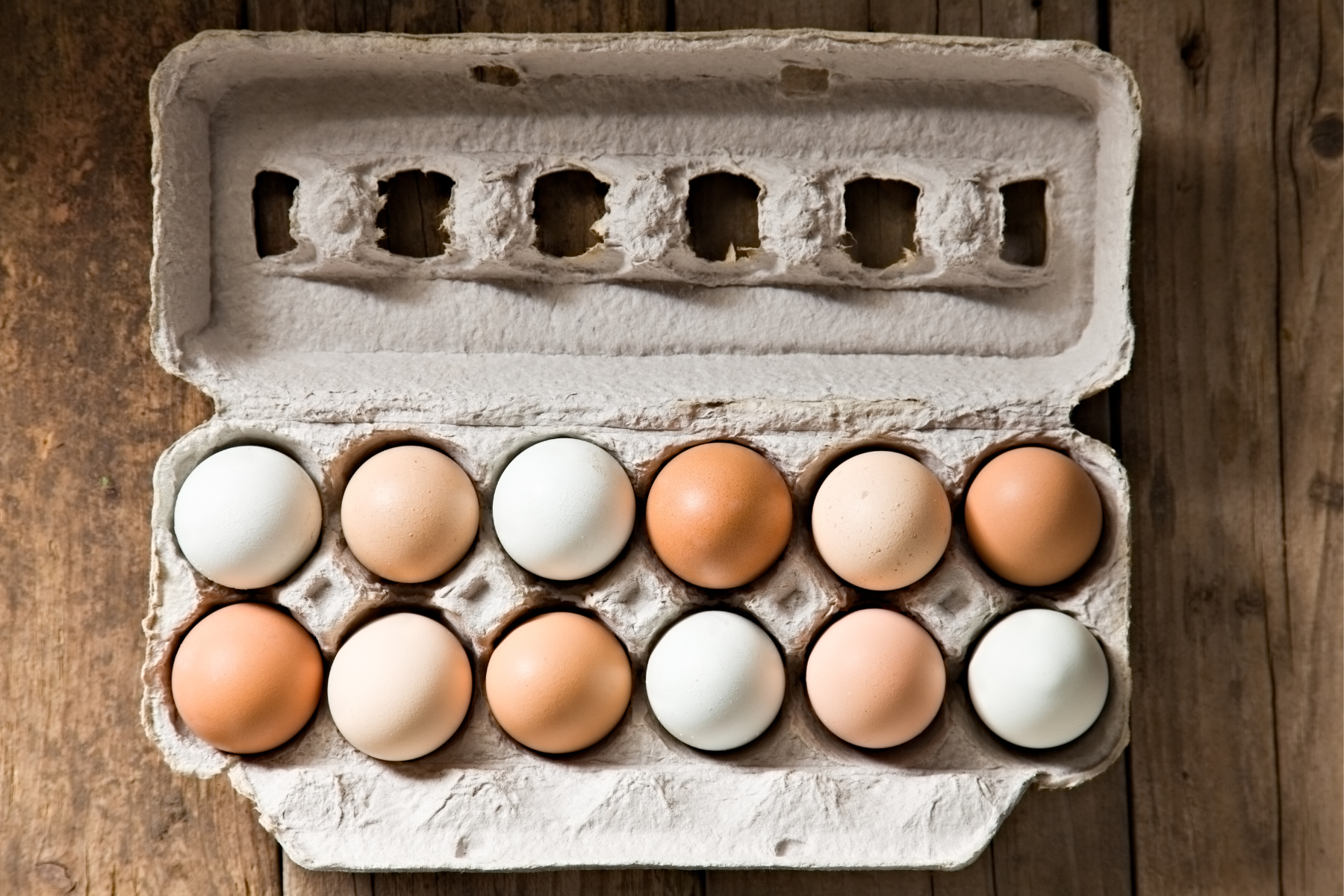 A dozen brown and white eggs sit in an open, grey cardboard carton on a rustic wooden table.