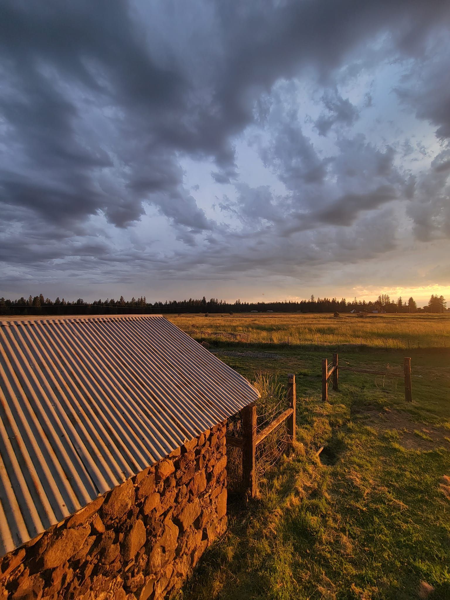 A sunset glows on a stone building with a corrugated metal roof, beside a grassy field under a dramatic, cloudy sky.