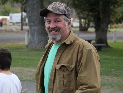 A smiling person in a camouflage cap and tan button-up shirt over a light green t-shirt, standing in a grassy park.