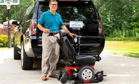 Man standing by a black SUV, loading a red lawn mower onto the rear lift at a driveway.