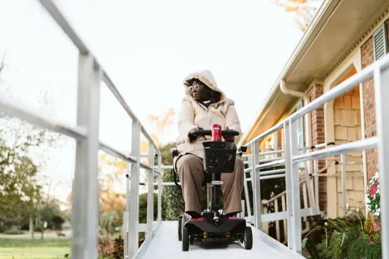 Person in a beige hooded jacket rides a mobility scooter up an outdoor ramp beside a building