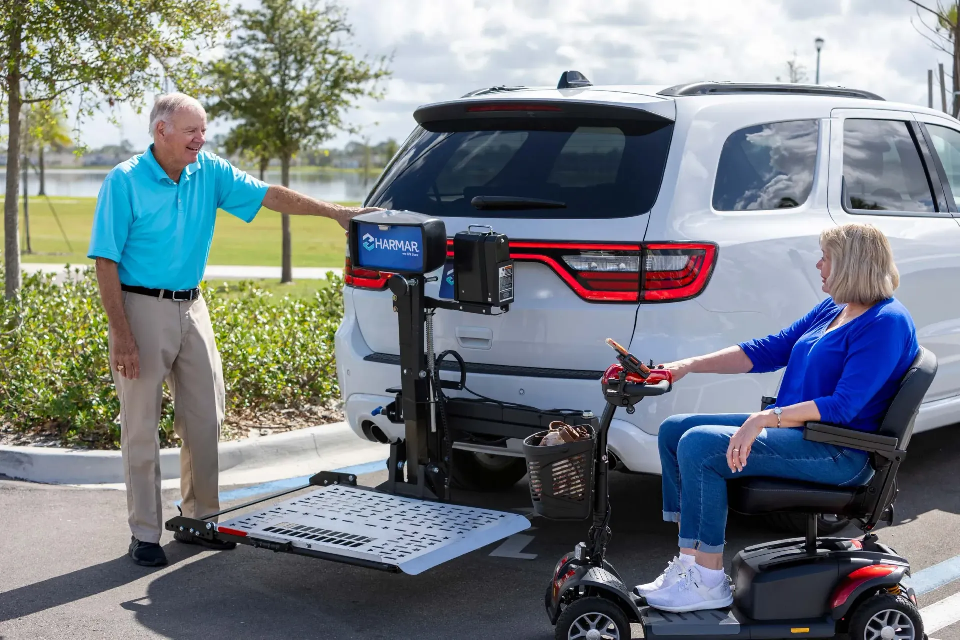 Man demonstrating a wheelchair lift at the rear of a silver SUV beside a seated person on a mobility scooter