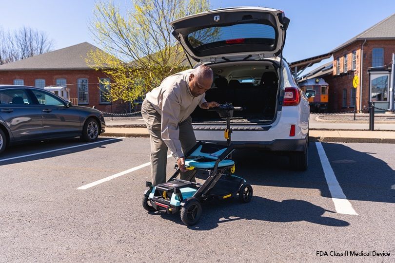 Man loading a folded mobility scooter into the back of an SUV in a parking lot