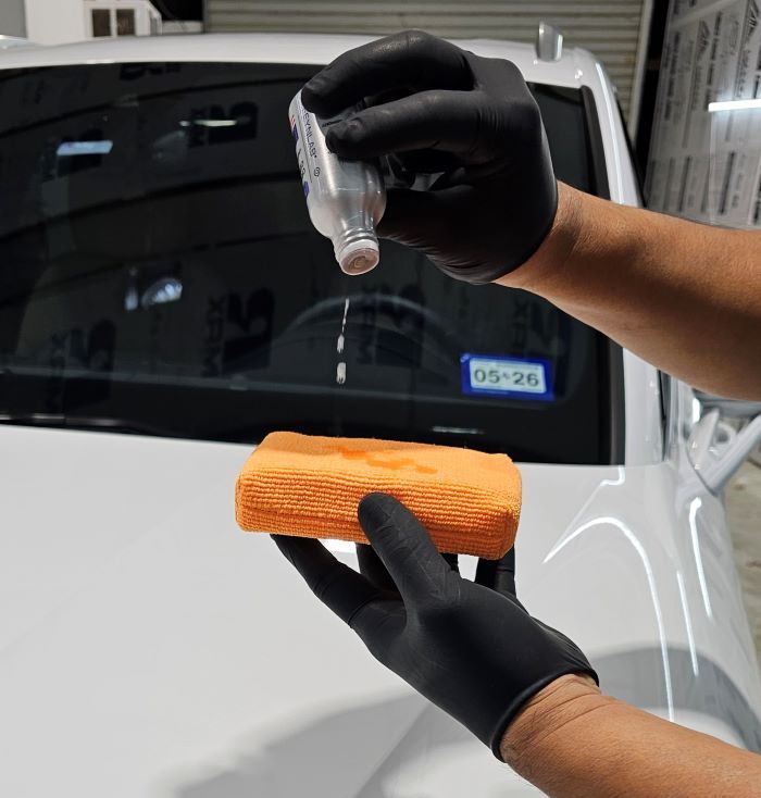 Hands in black gloves applying liquid to a car with a microfiber cloth.