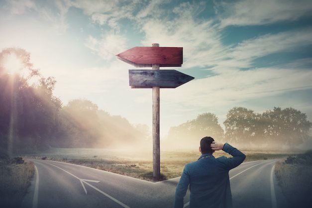 Person at a fork in the road, looking at a signpost in a misty landscape at sunrise.