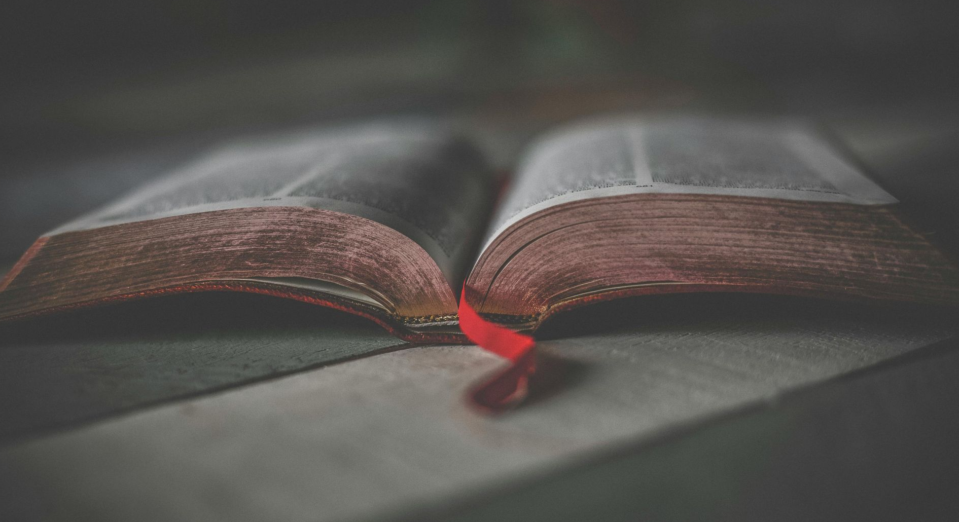 Open book with red ribbon bookmark on a gray surface, softly lit from above