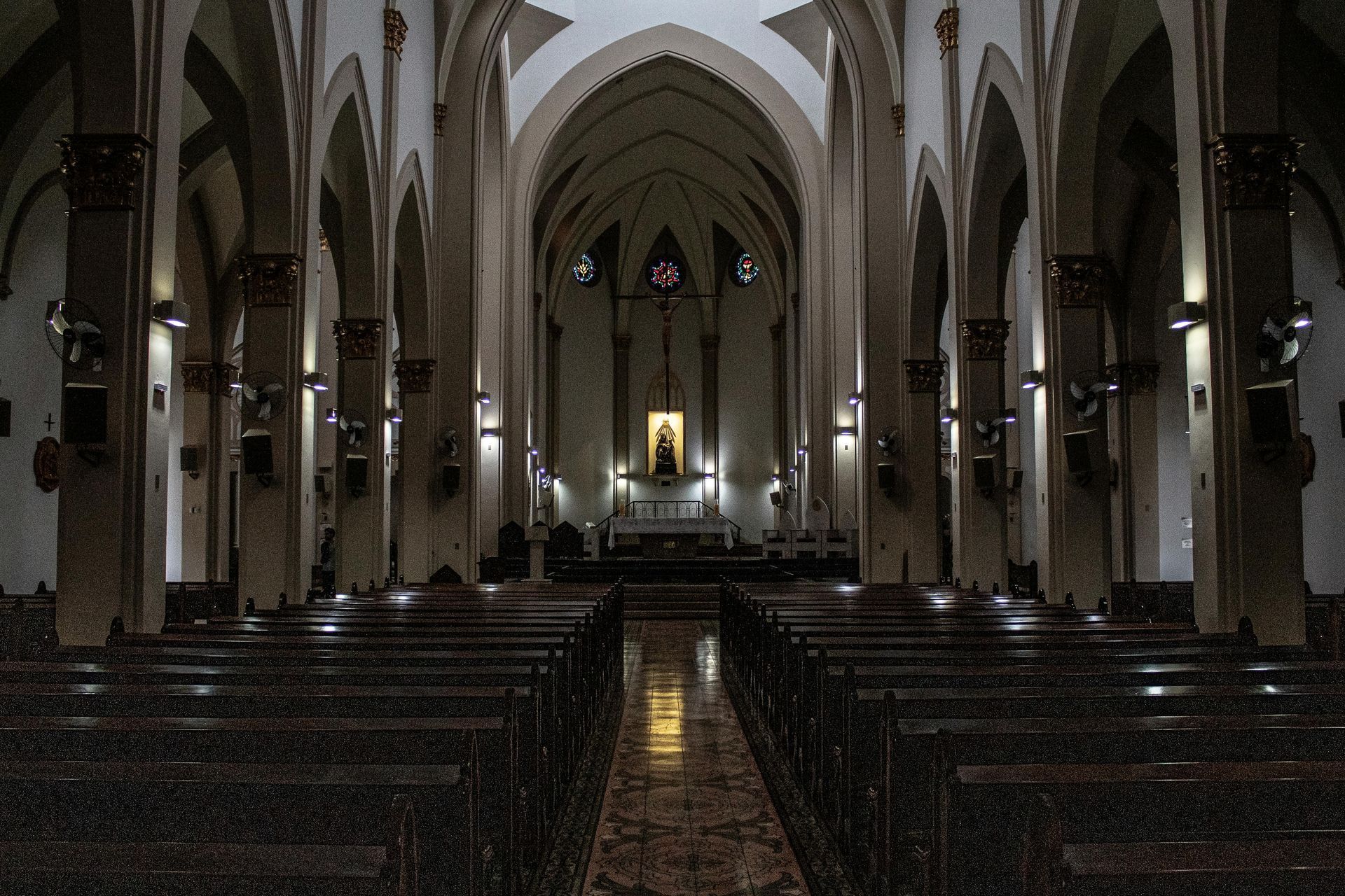 Dim cathedral interior with rows of pews and tall arches leading to a lit altar.