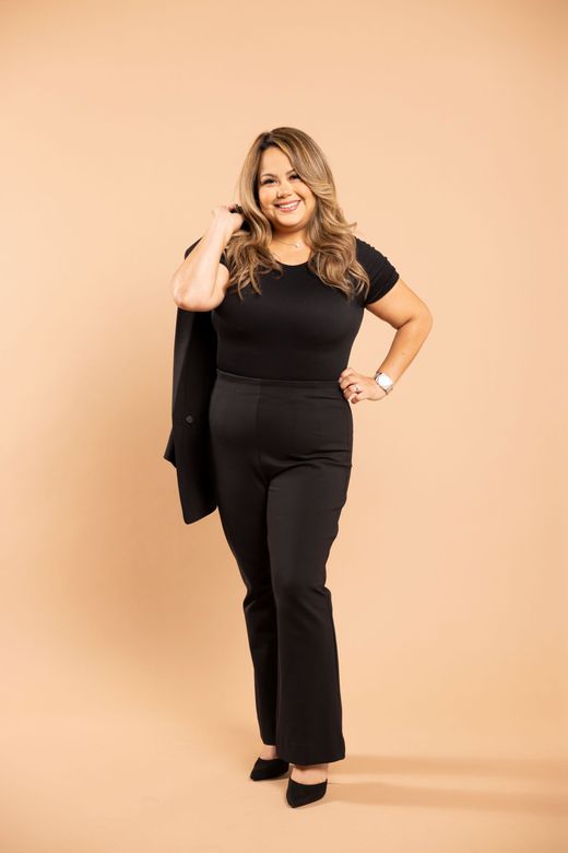Woman in black outfit posing against a peach studio backdrop, smiling with one hand on her hip.