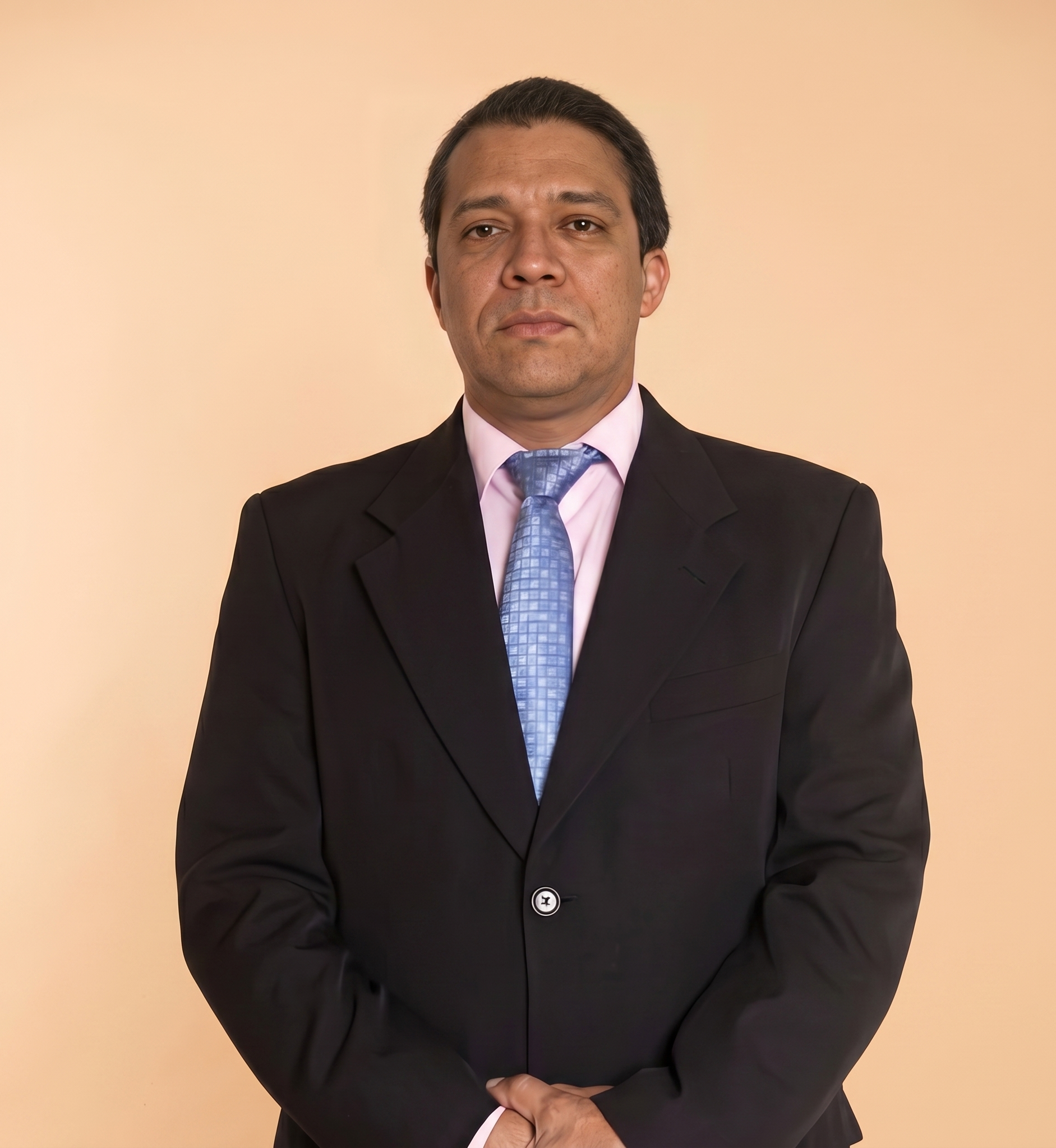 Man in a black suit and blue tie standing against a plain beige background, hands clasped.