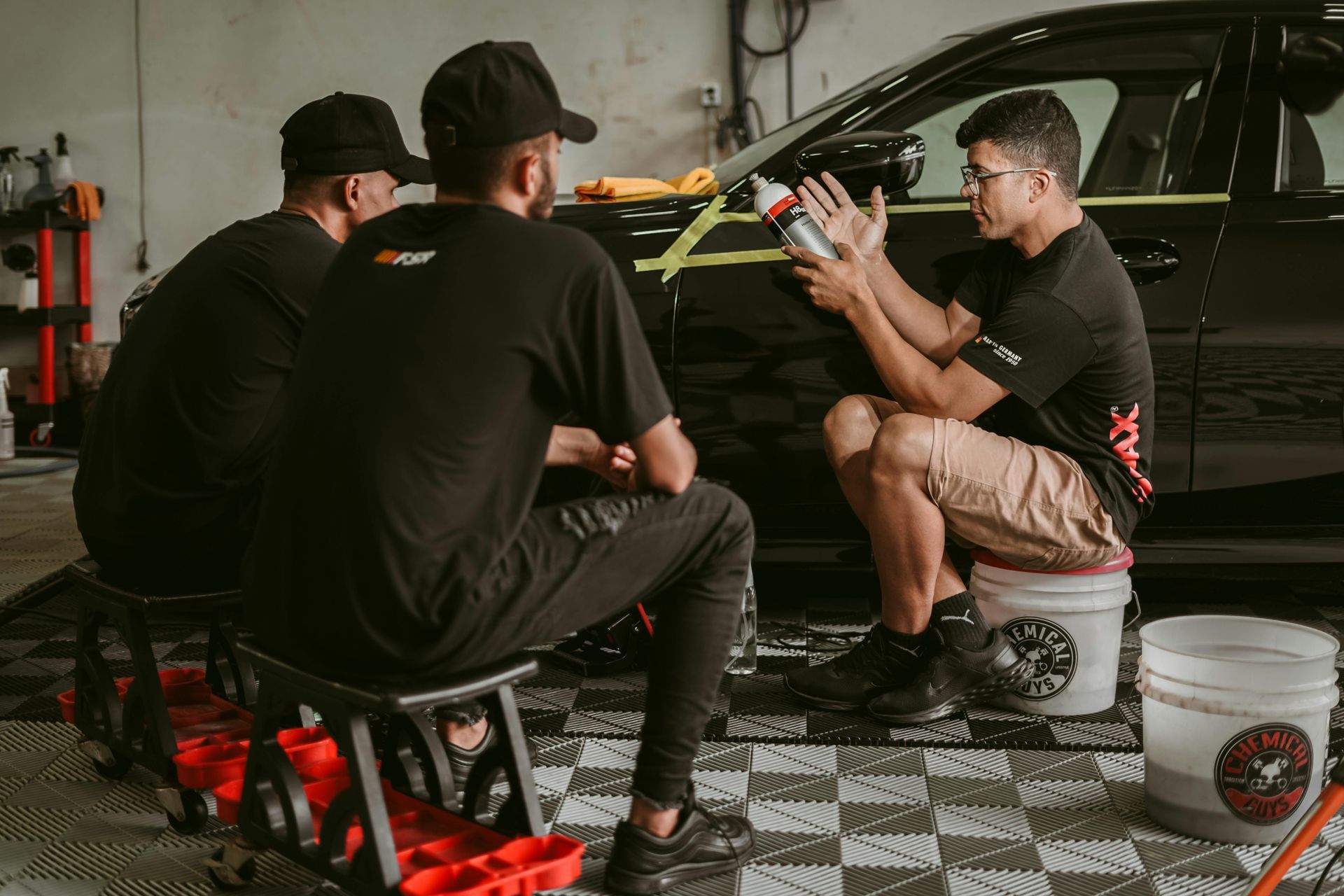 Three mechanics working on a black car in a garage, with tools and parts on the floor