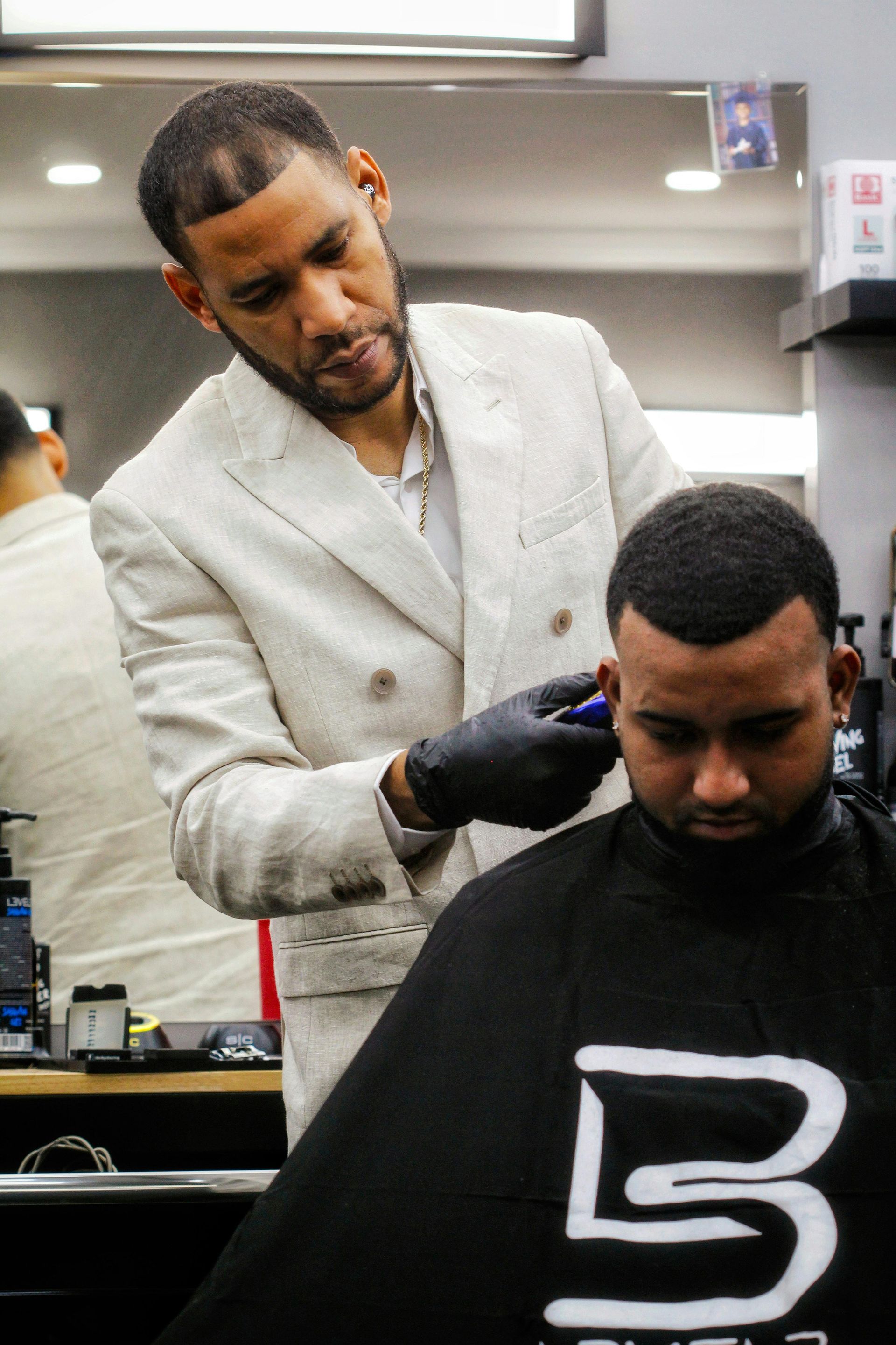 Barber in a light suit trims a seated client’s hair in a barbershop.