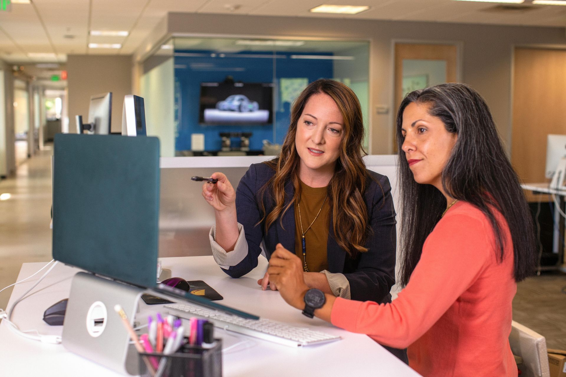 Two coworkers collaborate at a desk in a bright office, looking at a computer screen.