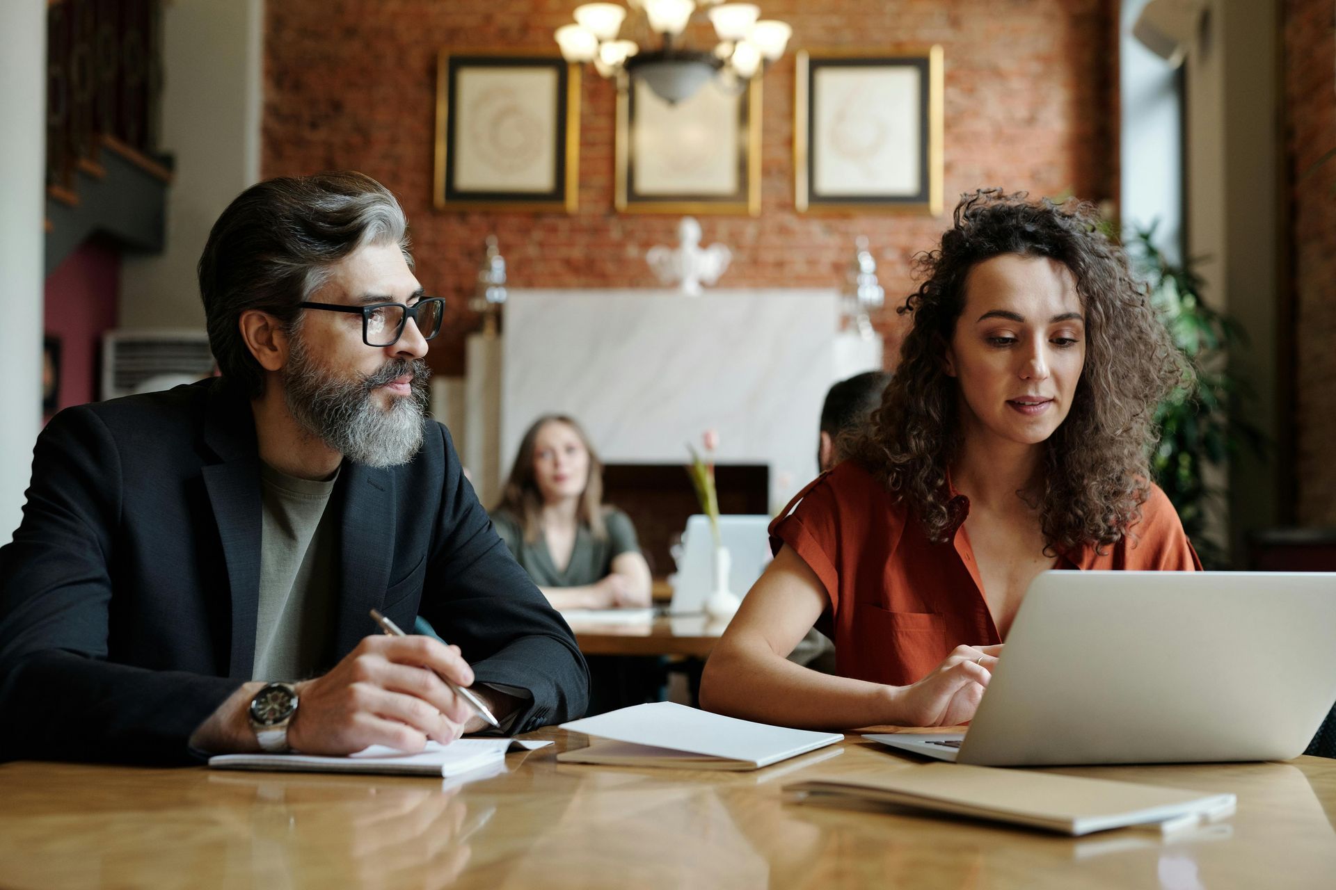 Two colleagues at a conference table, one taking notes and one using a laptop in a meeting room