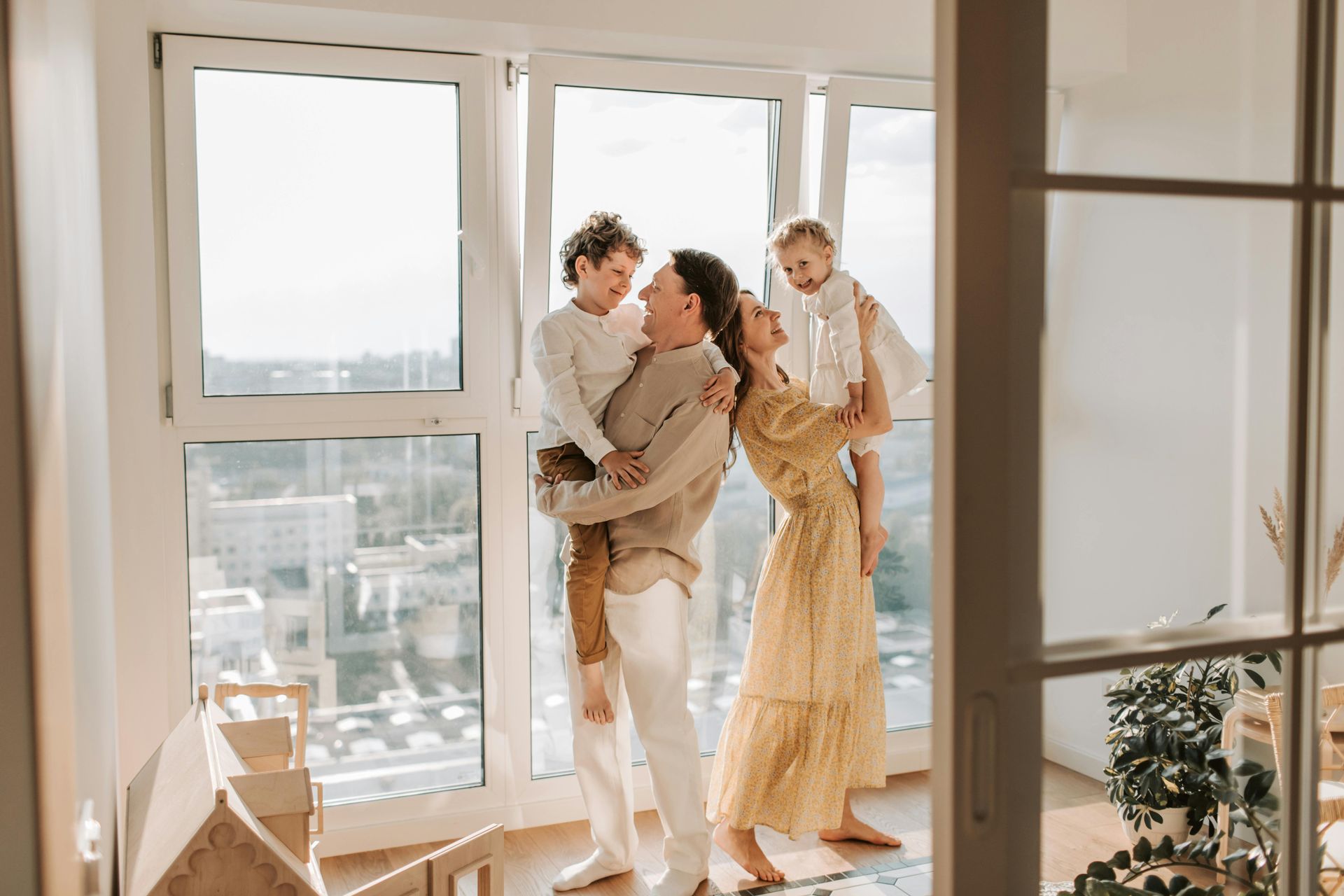 Two adults hold a child in a bright apartment by large windows, smiling in warm sunlight.