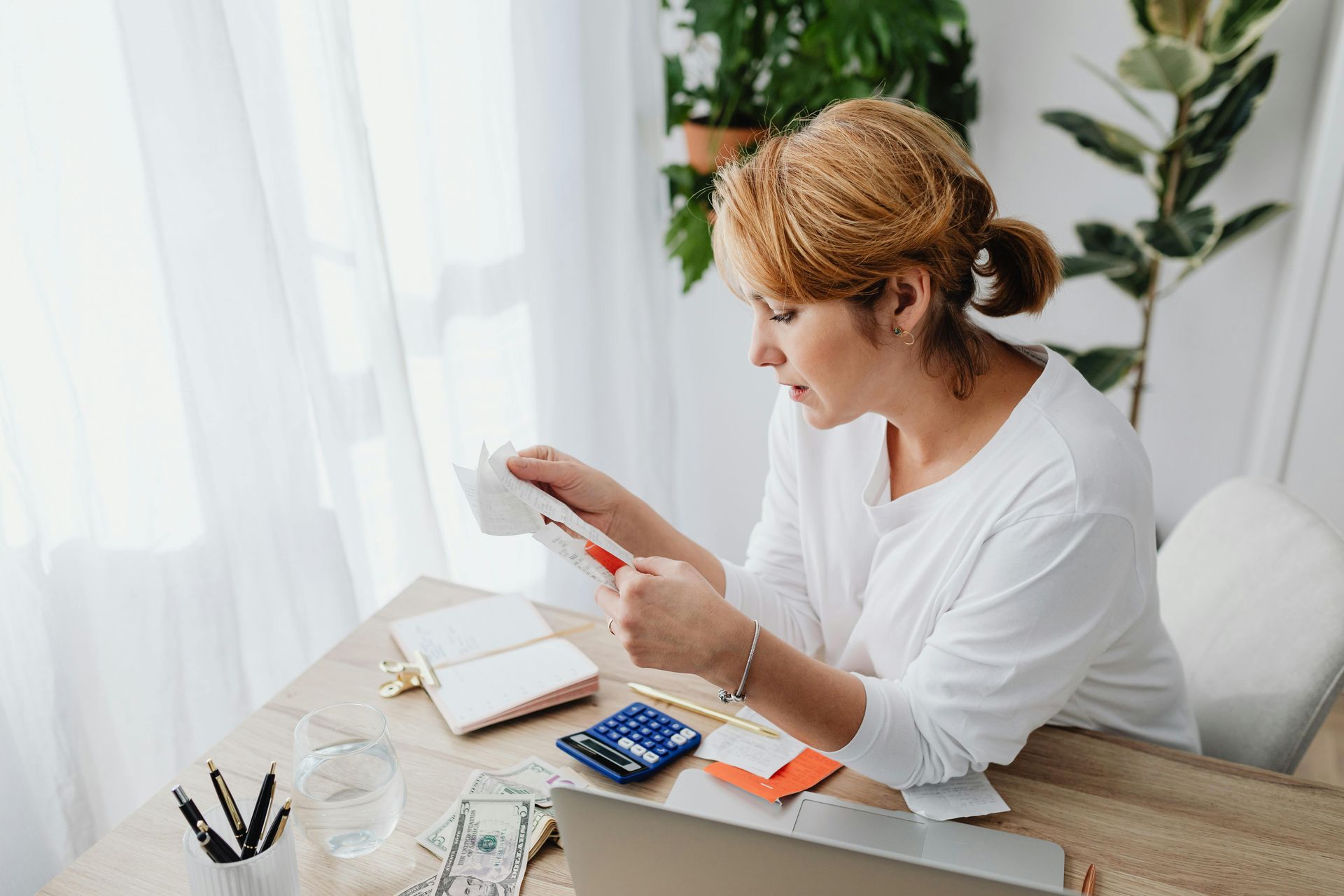 Woman at a desk using a smartphone beside a laptop and calculator in a bright home office