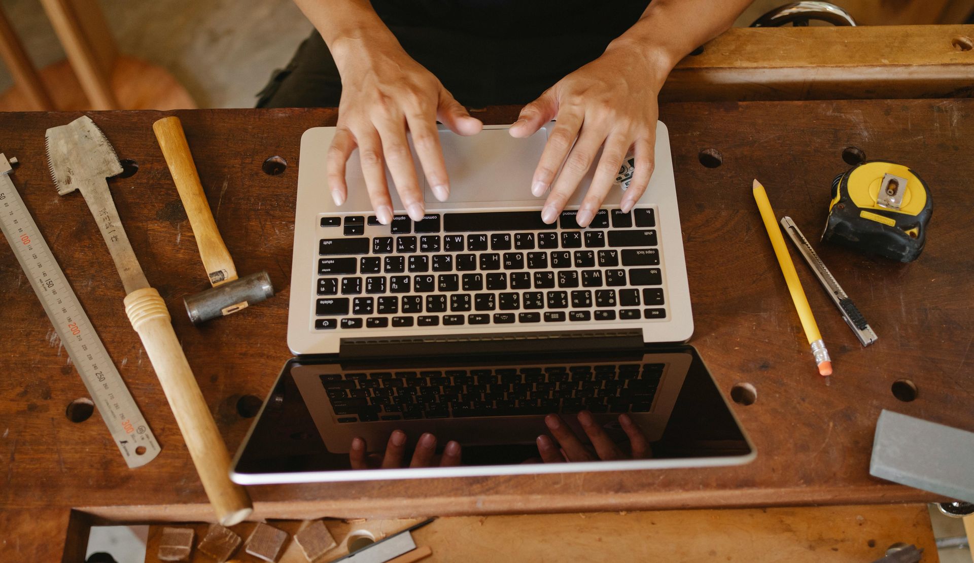 Hands typing on a laptop at a wooden workbench with tools, a tablet, and a tape measure nearby.
